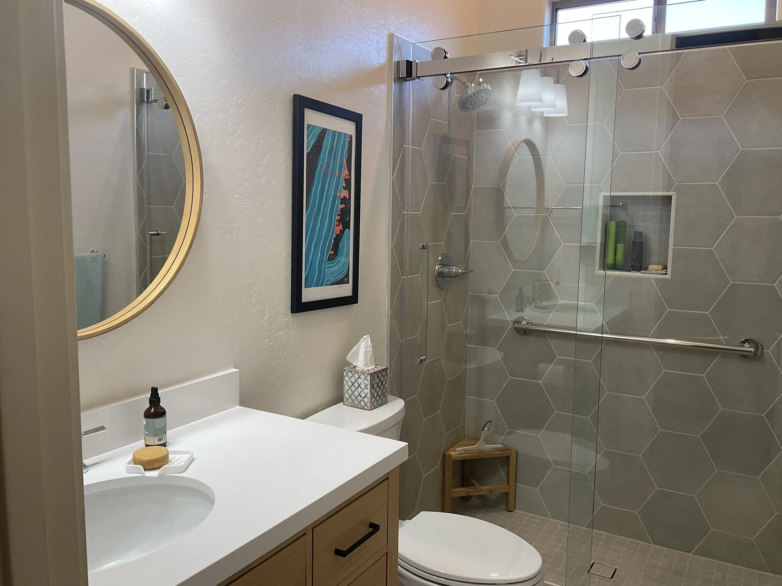 Bathroom with a vanity with a white countertop, a round mirror, soap and sanitizer on the counter, a tissue box, a framed artwork on the wall, a toilet, a small wooden stool, and a walk-in shower with beige hexagonal tiles, a built-in shelf with toiletries, a metal grab bar, and a glass door.