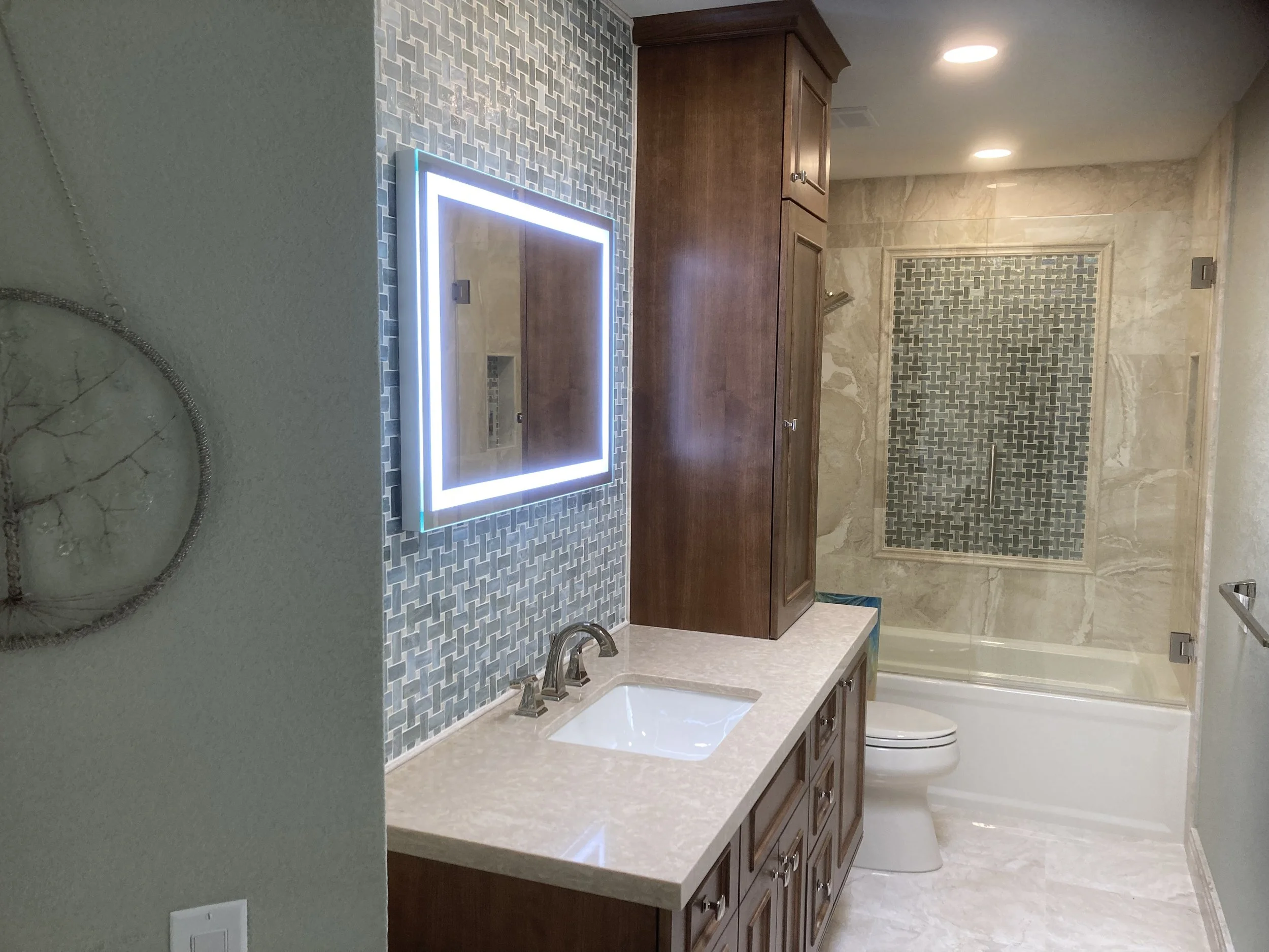 Bathroom with wooden cabinets, a lit mirror, a marble countertop with sink, tiled blue accent wall, a toilet, and a bathtub with a glass shower door featuring a decorative tile pattern.