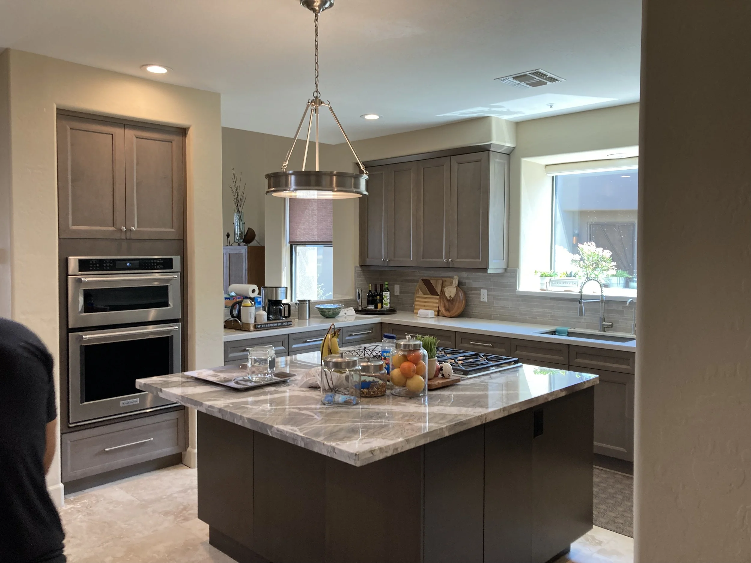 Modern kitchen with granite island, wooden cabinets, and stainless steel appliances illuminated by ceiling lights and sunlight from a window.