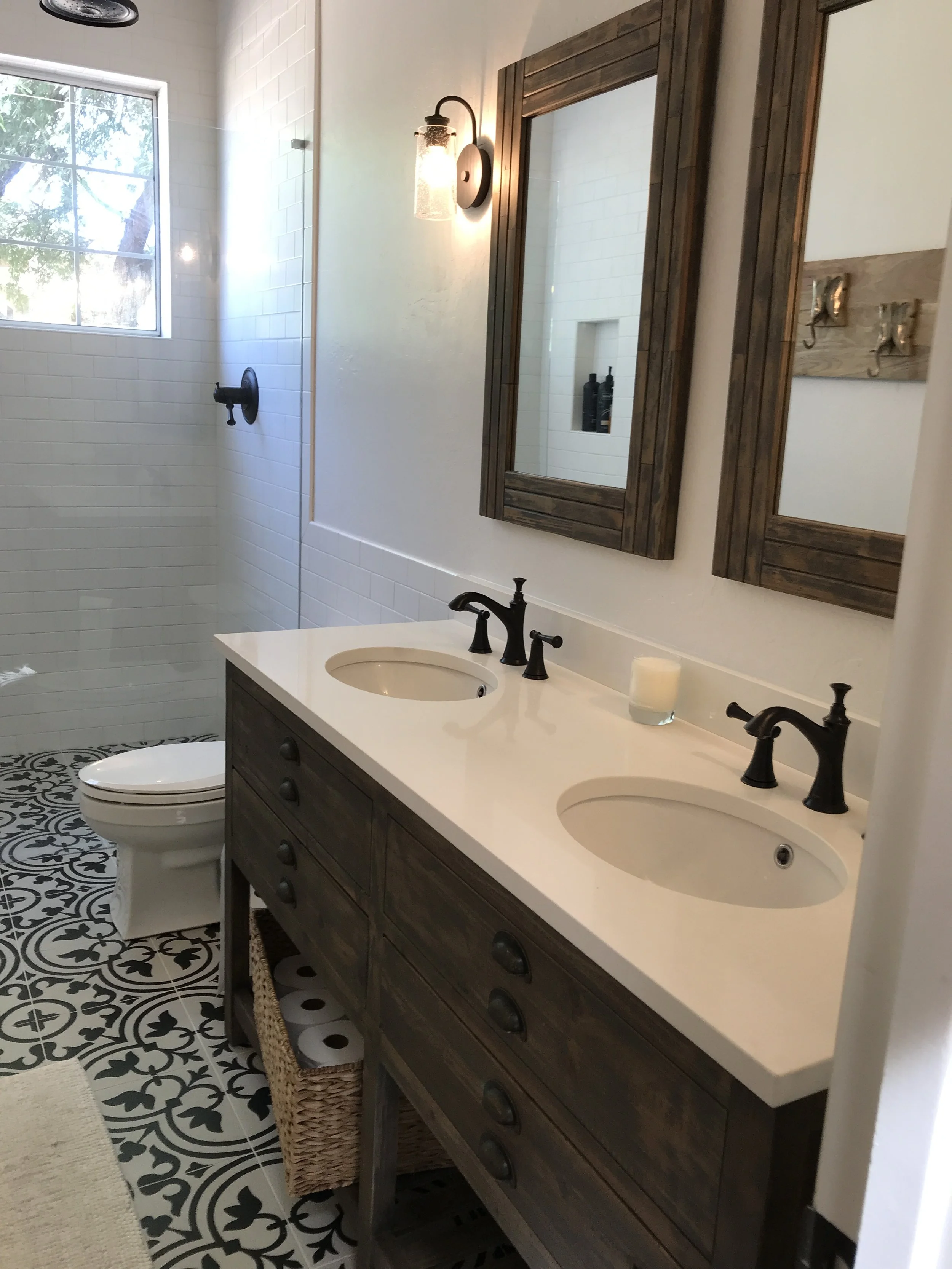 A bathroom with a dual sink vanity, wooden framed mirrors, black faucets, a white countertop, and a patterned tile floor. The shower area has white tiles, a window, and black fixtures.