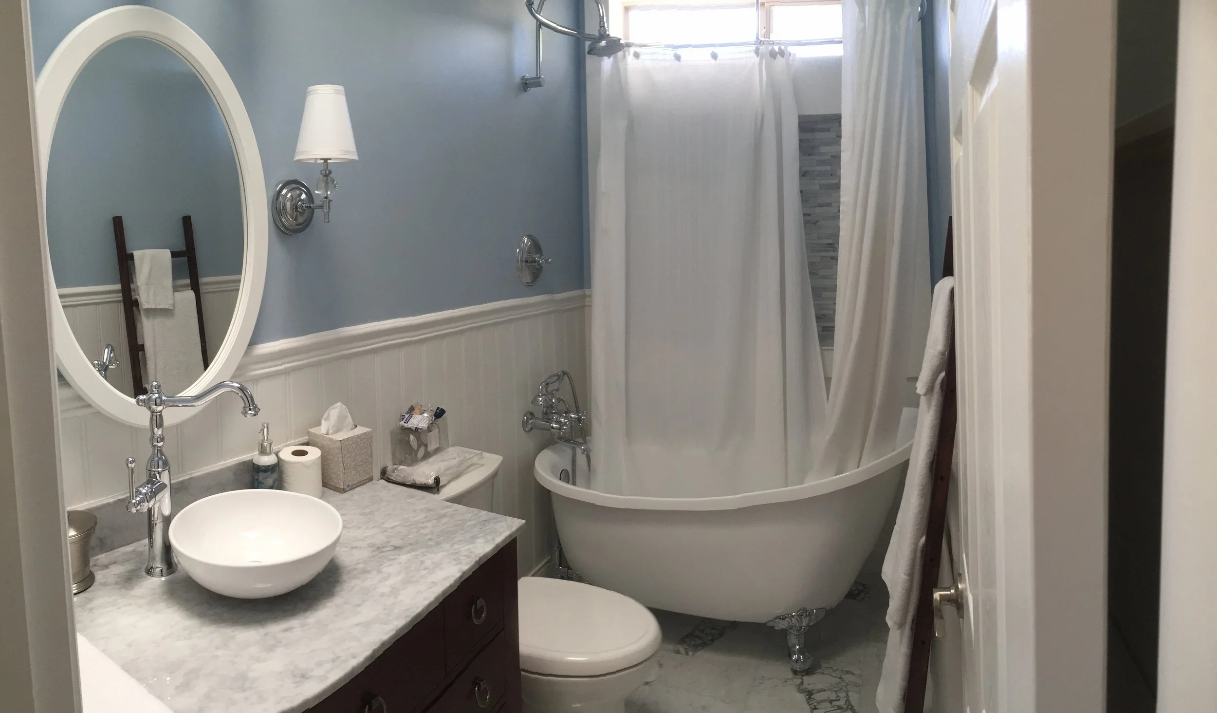 A bathroom with a blue upper wall and white wainscoting, featuring a white oval mirror, wall-mounted lamp, bathtub with a white curtain, marble countertop with a vessel sink, toilet, and a wooden towel ladder with towels.