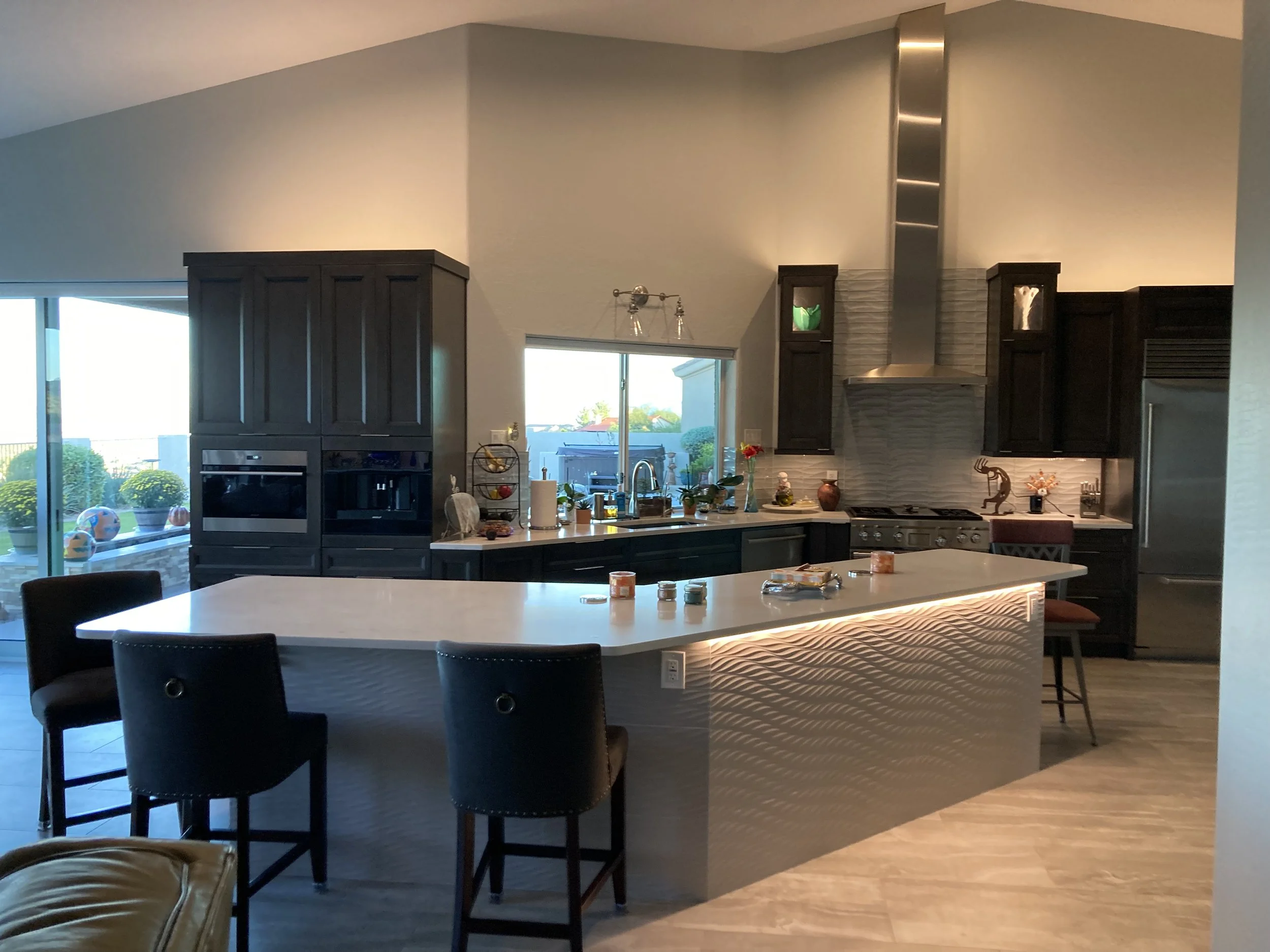 Modern kitchen with a large white island, dark cabinetry, stainless steel appliances, and a window overlooking a patio with potted plants.