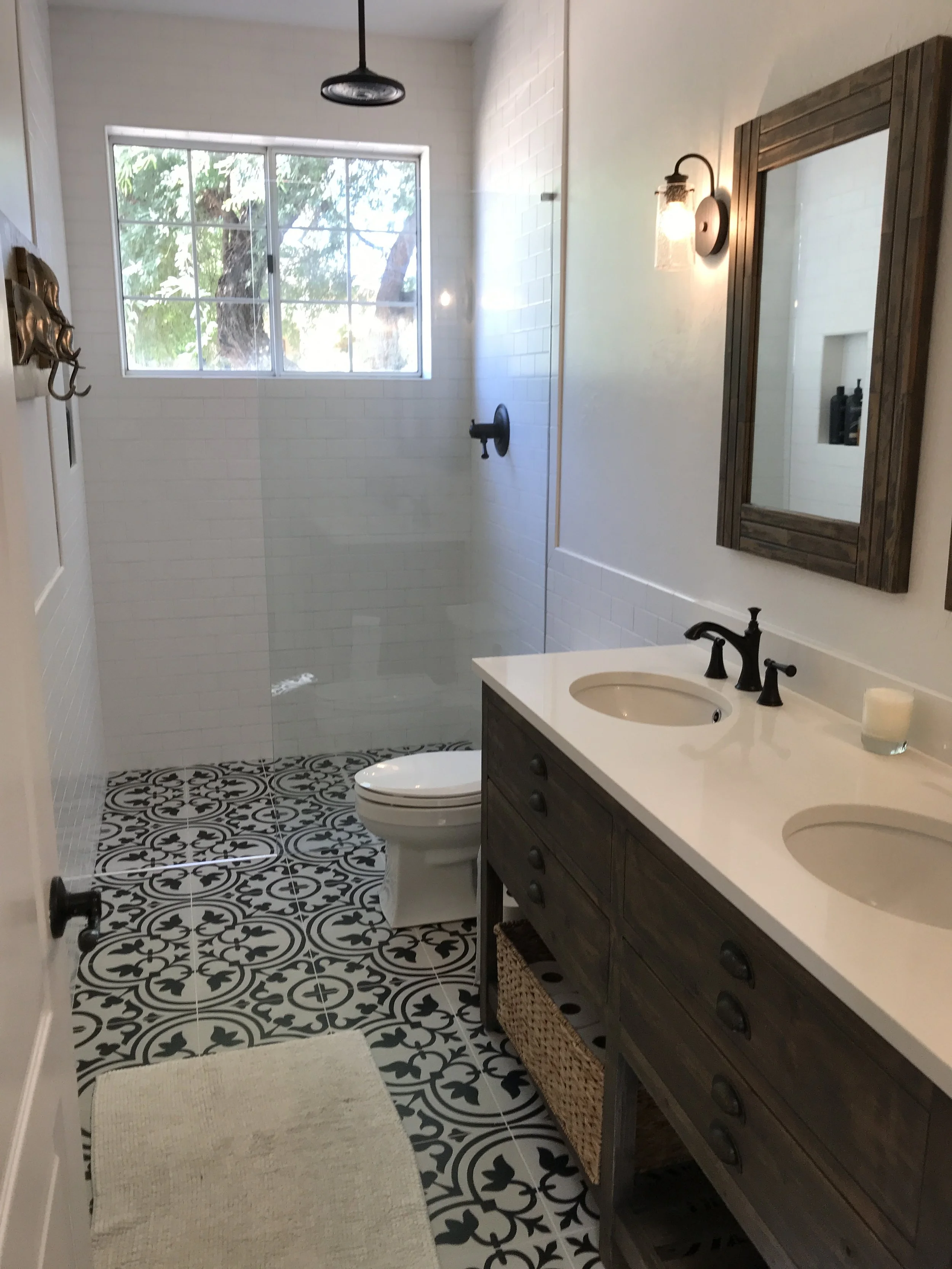 Bathroom with double vanity, white countertop, black fixtures, large mirror, walk-in shower with black fixtures and a window, patterned black and white floor tiles, beige rug, and wall-mounted light fixtures.