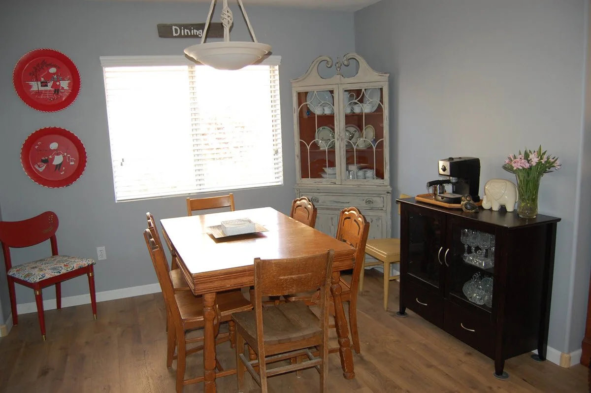 A dining room with a wooden table and chairs, a white china cabinet, a black sideboard with a coffee maker, a vase of pink flowers, and red decorative plates on the wall.