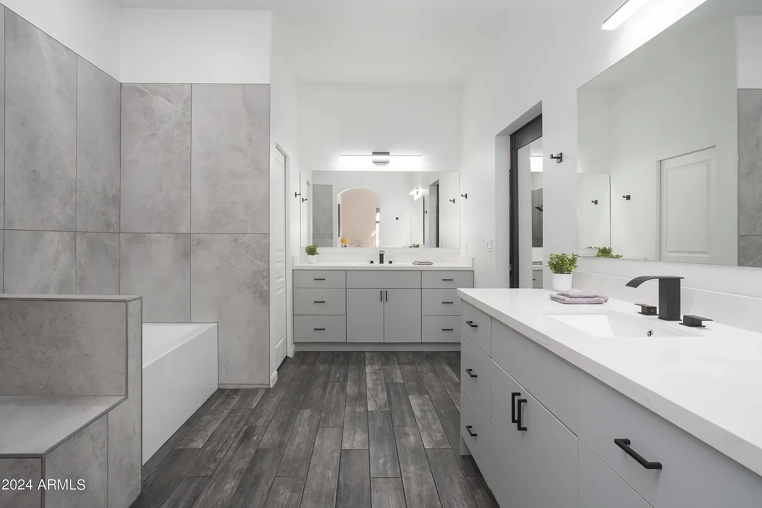 Modern bathroom with white cabinets, gray tiled walls, and dark wood flooring. Includes a large mirror, two small potted plants, and a black faucet.