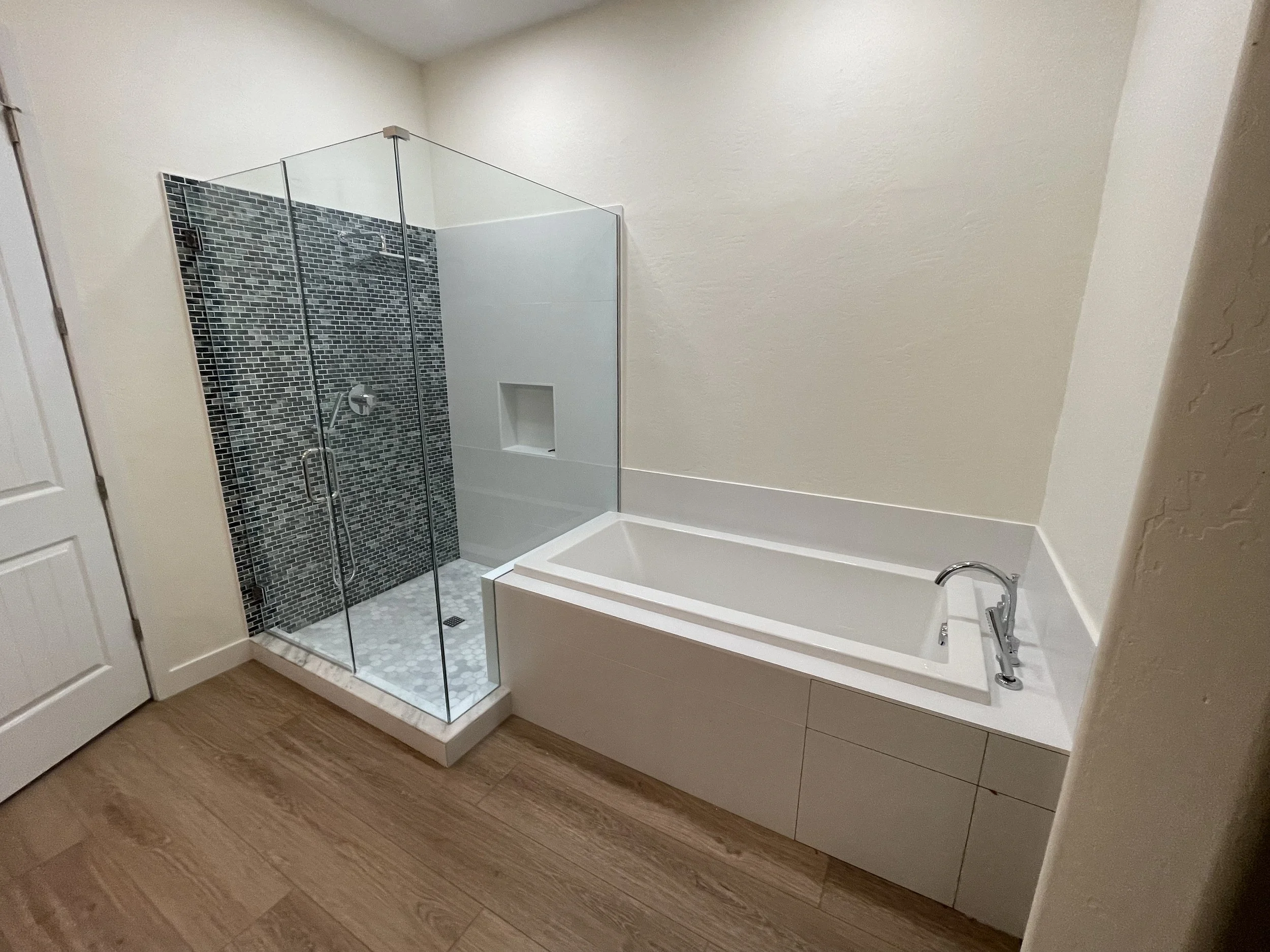 Bathroom with a glass-enclosed shower featuring dark mosaic tiles and a white bathtub with a chrome faucet on a wooden floor.