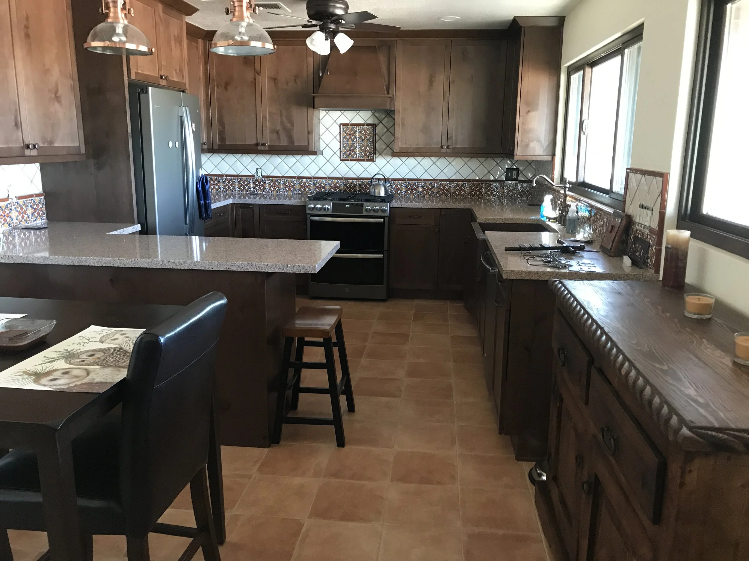 A kitchen with wooden cabinets, beige countertops, and a mosaic tile backsplash. There is a stainless steel refrigerator, a black oven with a stove, and large windows letting in natural light. A space with candles and a wooden countertop is on the ri