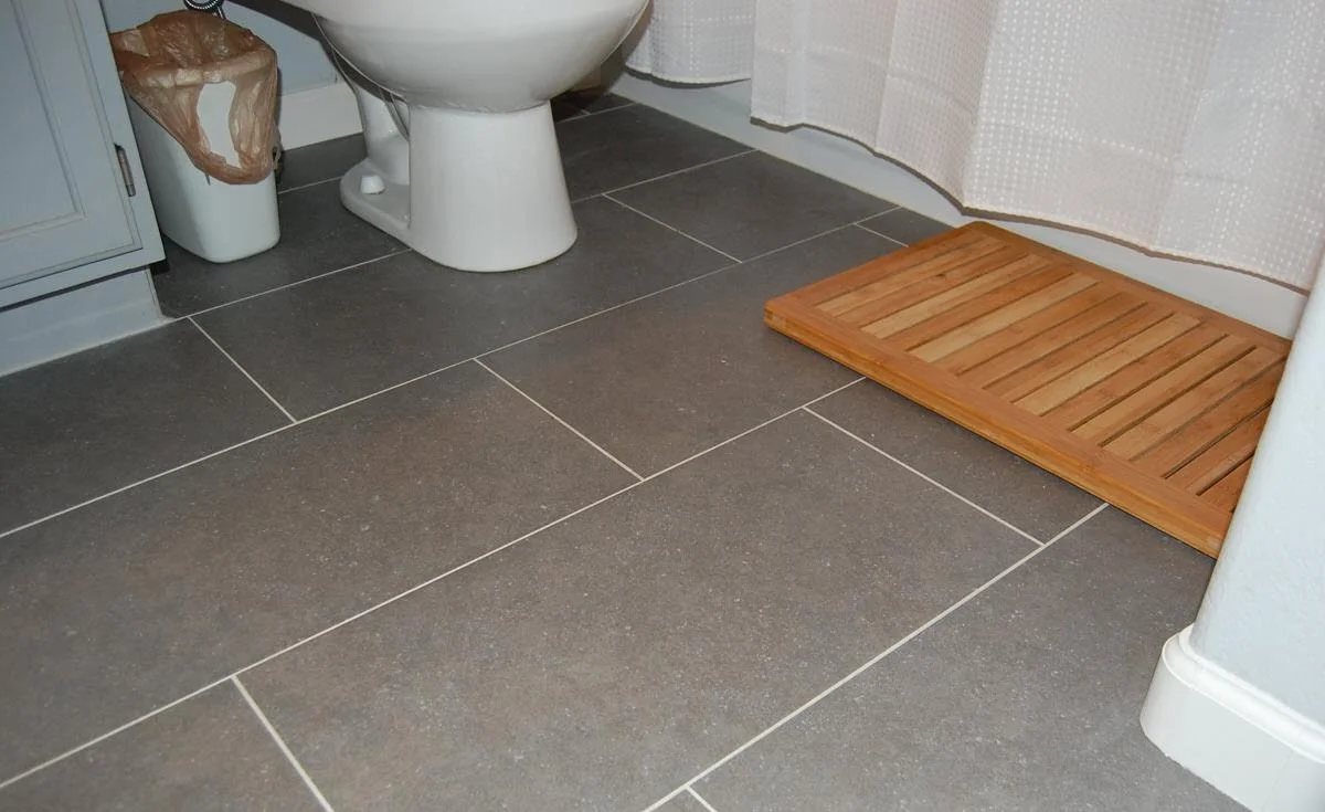 Bathroom floor with grey tiles, a white toilet, a small trash bin, a wooden bathroom mat, and a shower curtain.