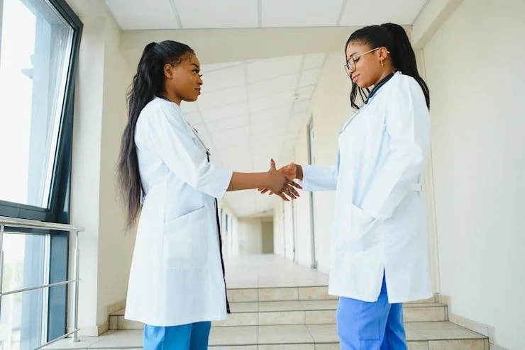 Two young women in medical uniforms shaking hands in a hospital corridor.