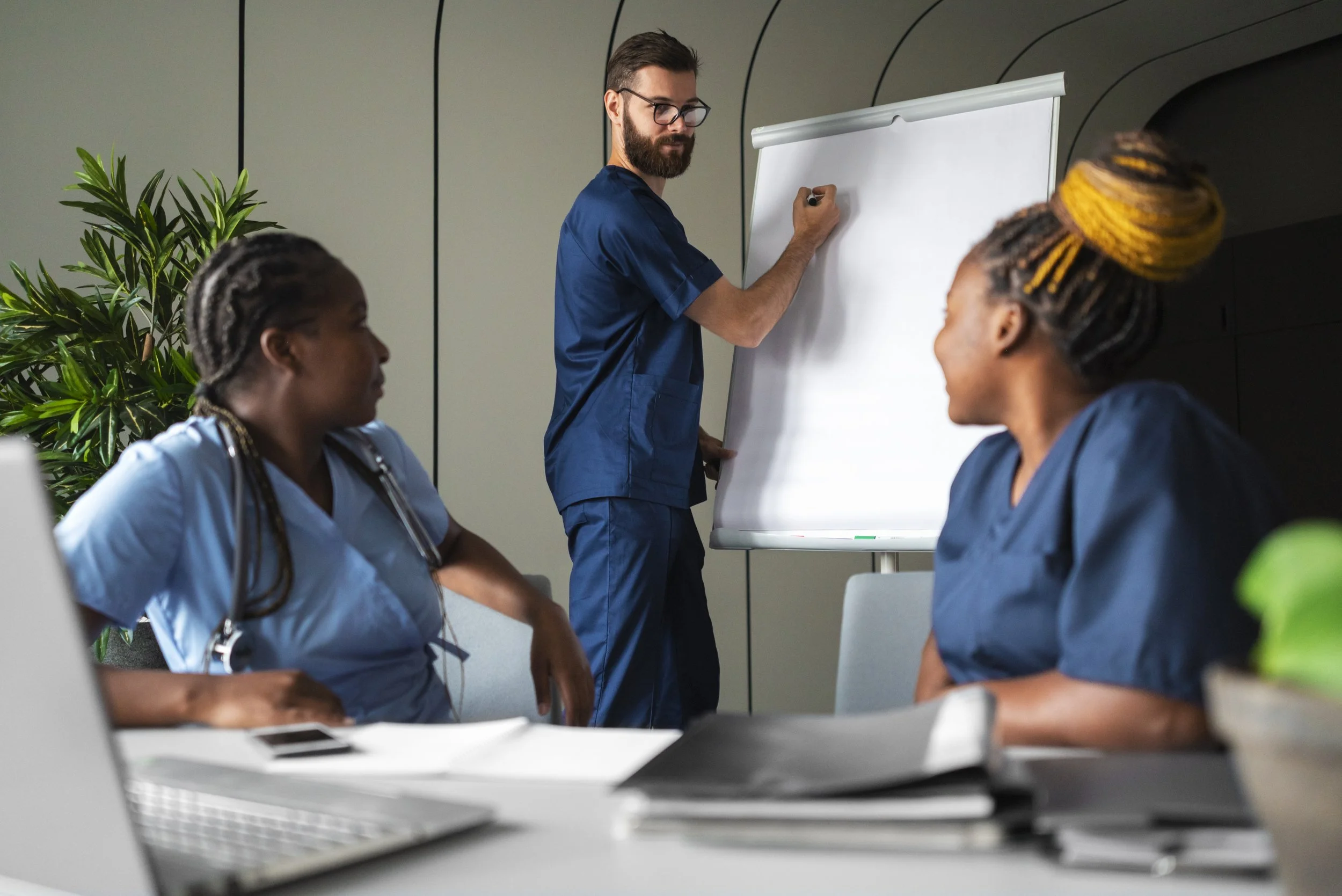 A male healthcare professional writing on a whiteboard in a meeting room with two female nurses seated at a table, with a laptop and documents on the table and a green plant in the background.