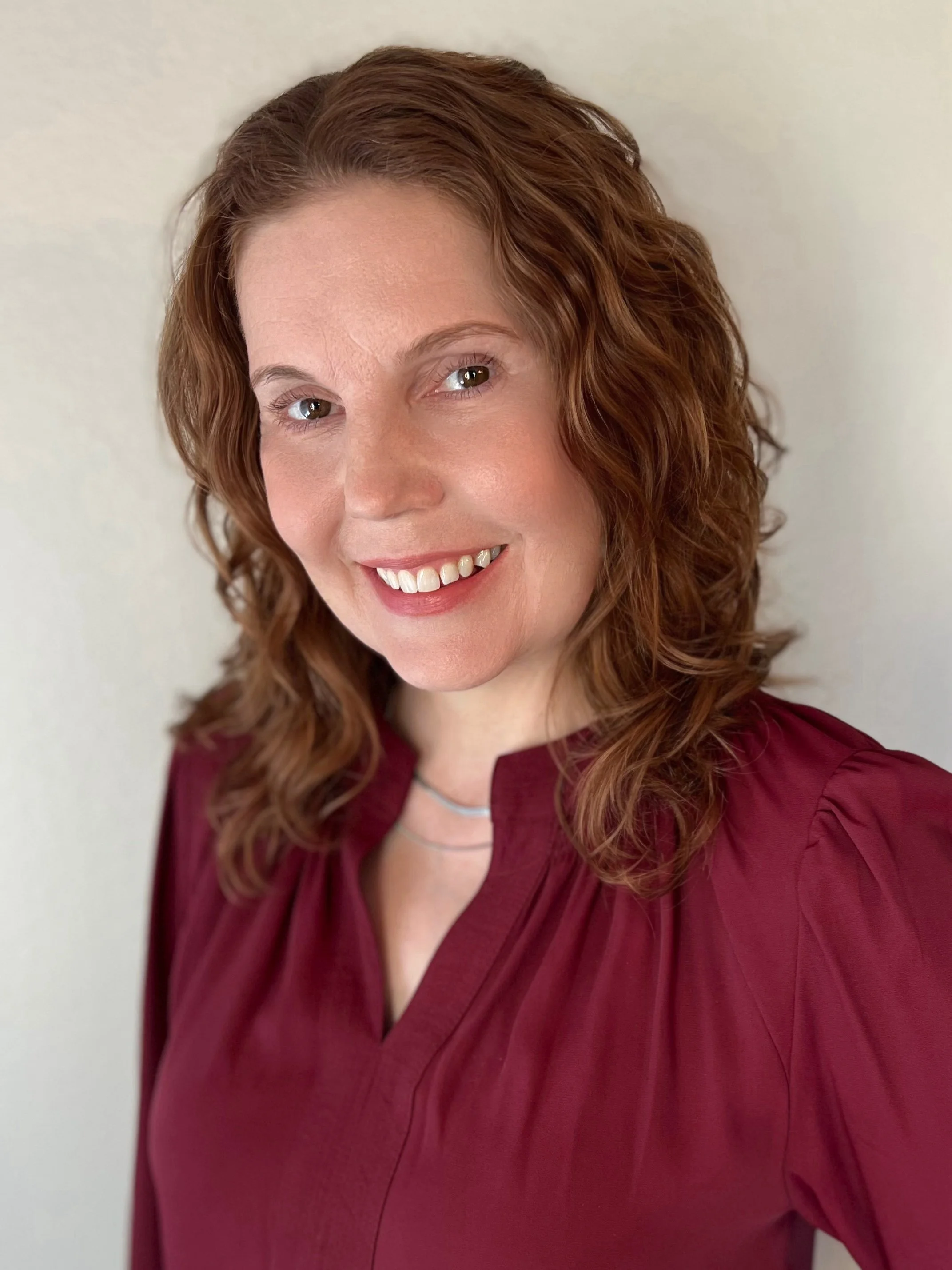 A young woman with shoulder-length auburn hair, smiling with her arms crossed, wearing a black knitted cardigan over a white top, standing in front of a plain light-colored background.