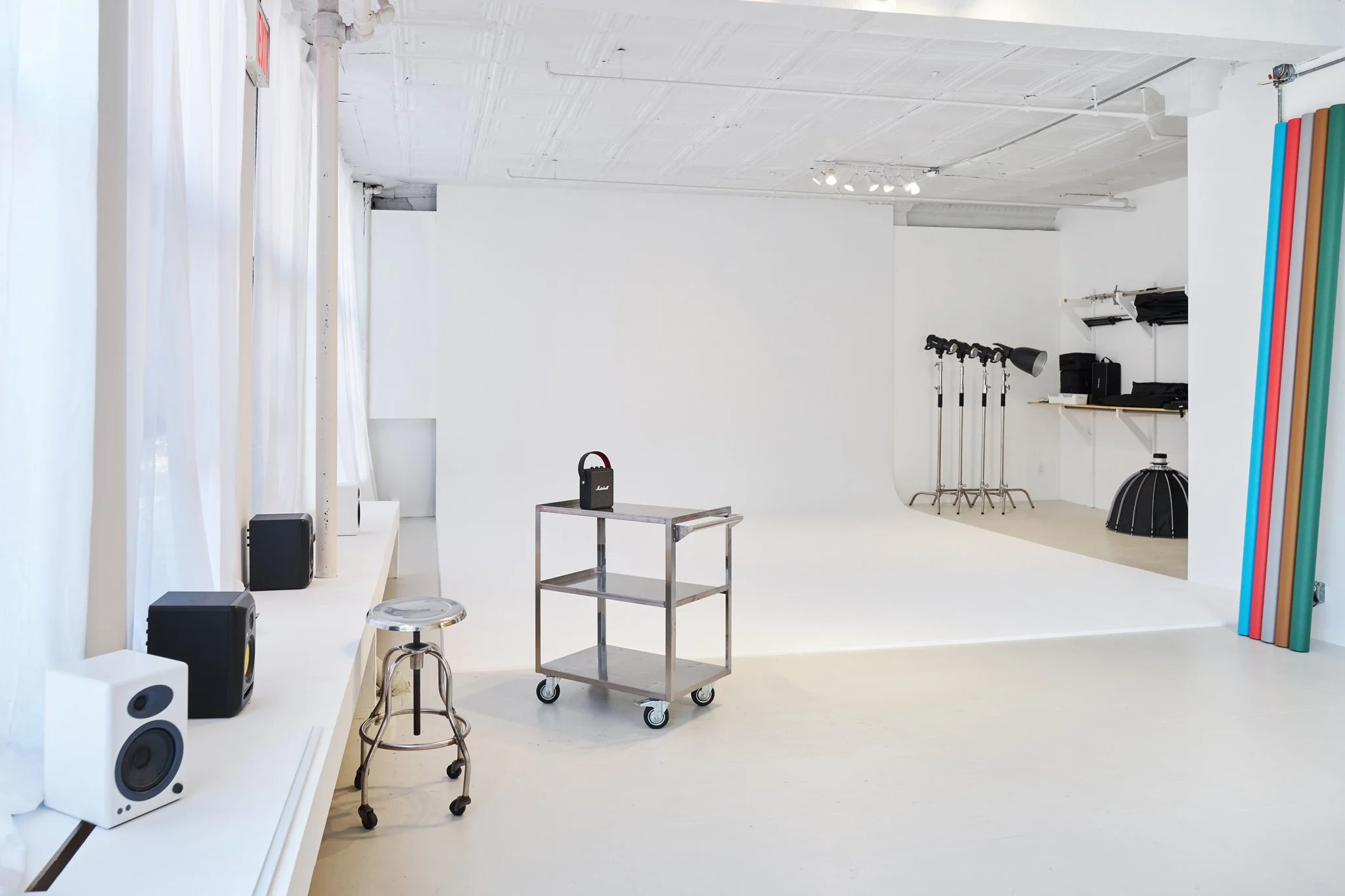 A bright, minimalist photography studio with a white backdrop, various lights, and equipment stored on shelves and the floor.