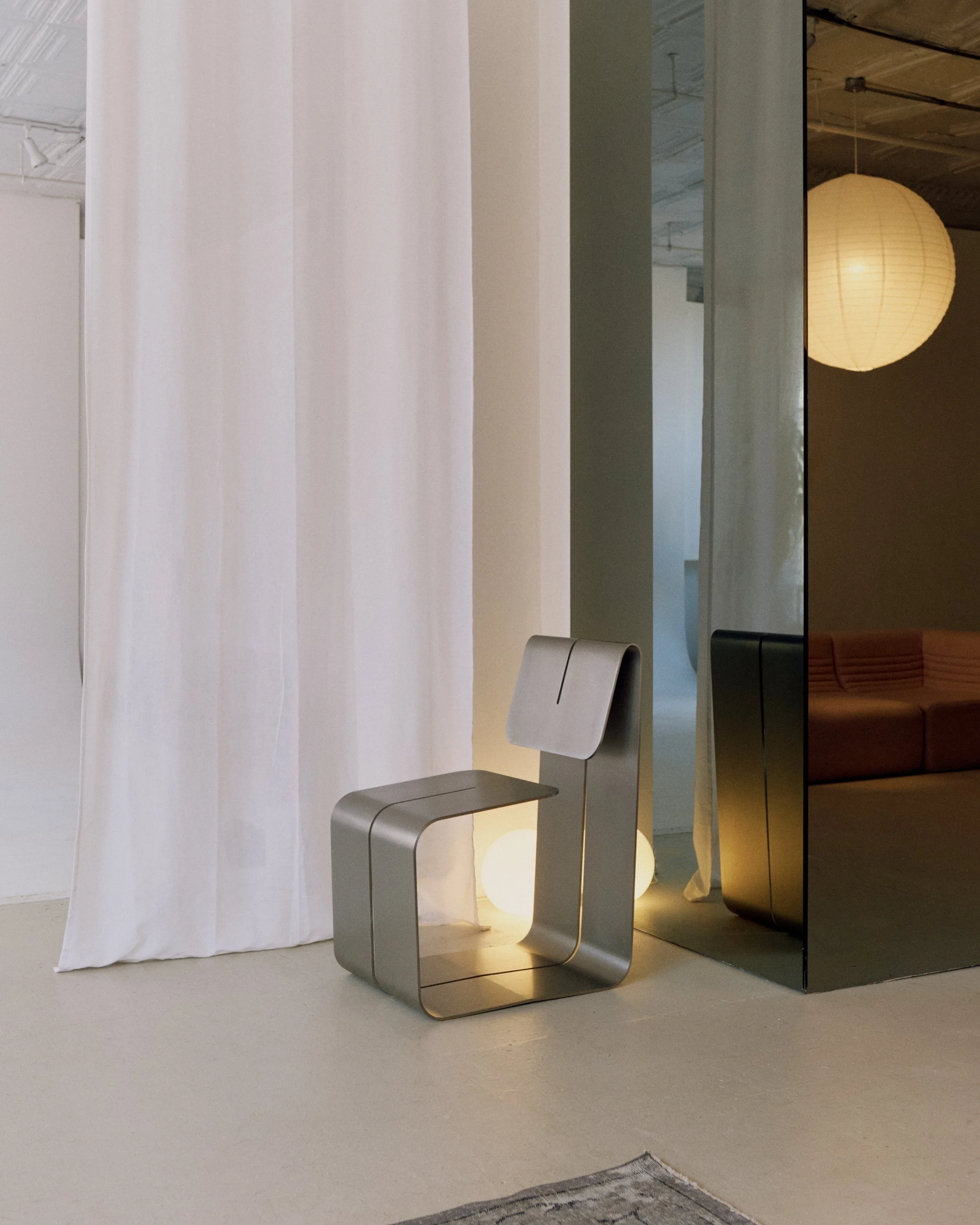 A modern metallic chair with a unique square-shaped backrest, placed in front of a white curtain. A soft light emanates from behind the chair, creating a warm glow. In the background, part of a workspace or showroom is visible, with a sofa and a paper lantern hanging from the ceiling.