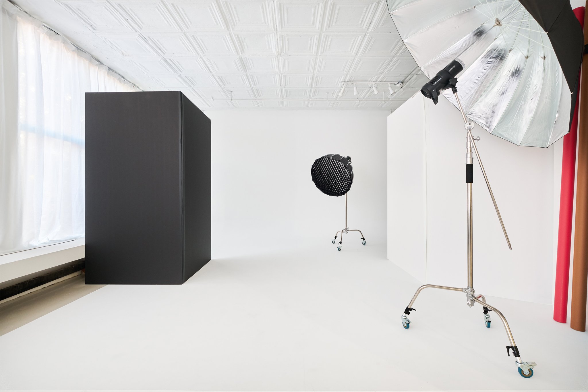 Photography studio with white background, black curtain, large black box, professional lighting equipment, including a softbox and a reflector, with colored rolls of seamless paper in the corner.