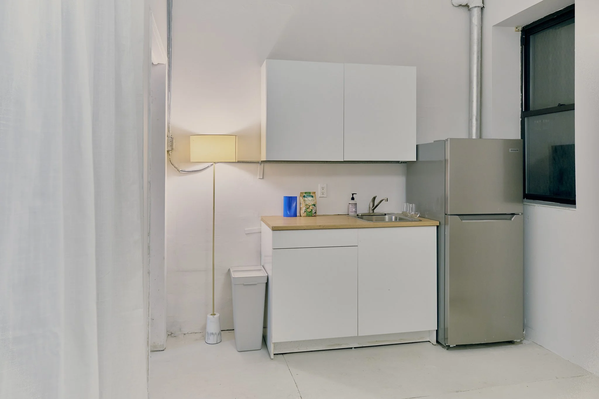 Small kitchen with white cabinets, a small refrigerator, a sink with a soap dispenser, a trash can, a blue cup, a box of cereal, a lamp, and a window.