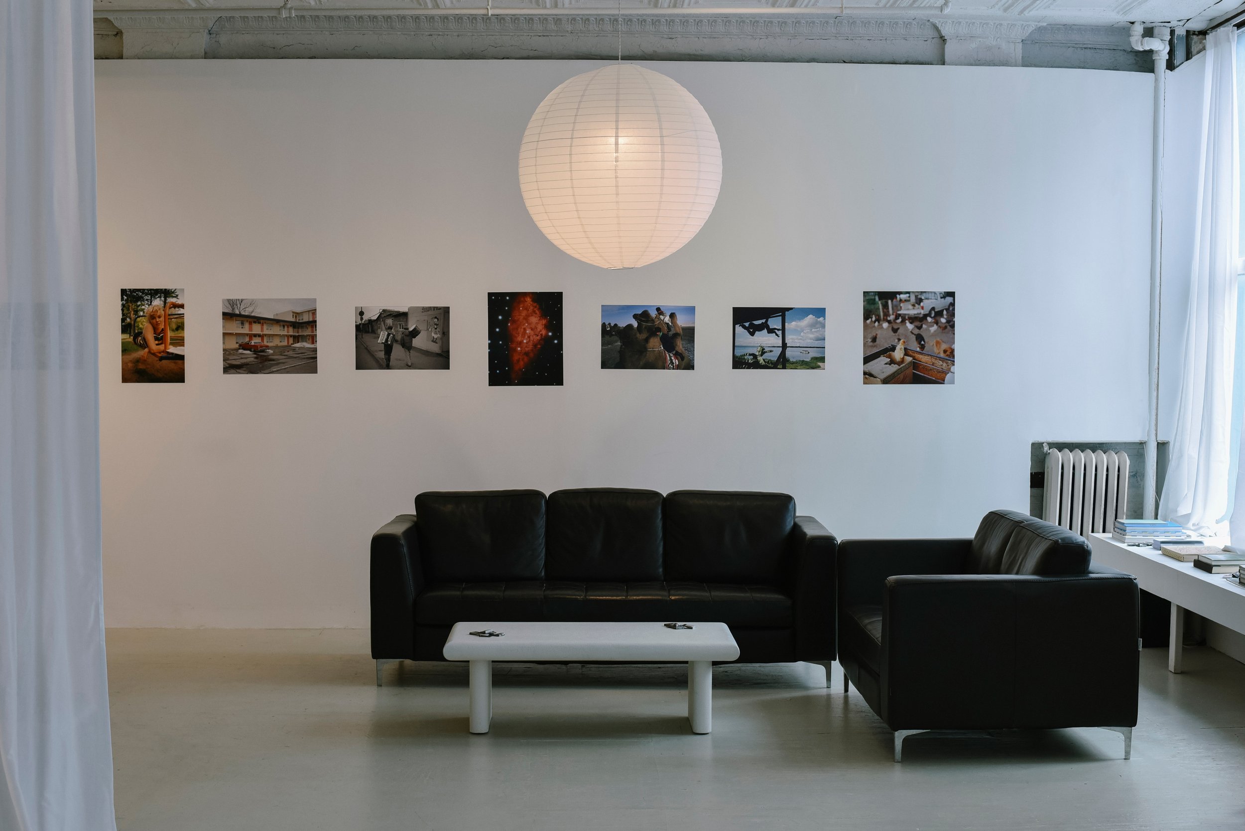 An art gallery interior featuring a white wall with seven framed photographs, a large white paper lantern hanging from the ceiling, a black leather sofa set, a small white coffee table, and a window with sheer white curtains letting in natural light.
