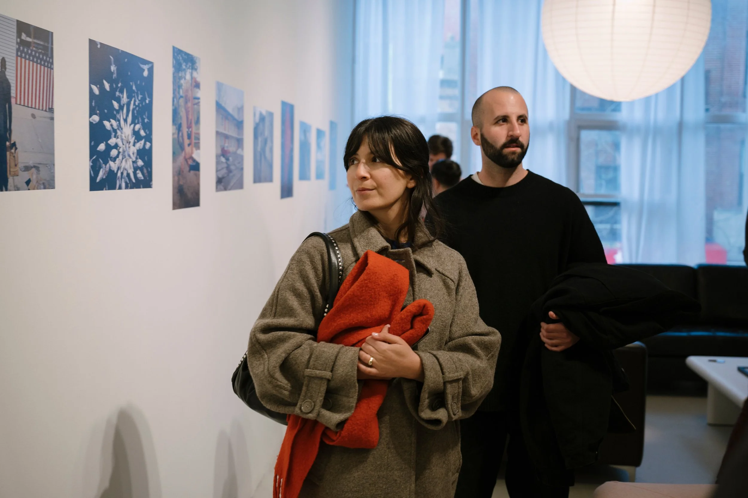 People observing artwork in a gallery, woman with dark hair holding a red coat, man with a beard, bald, wearing a black shirt, looking at the art on the wall.