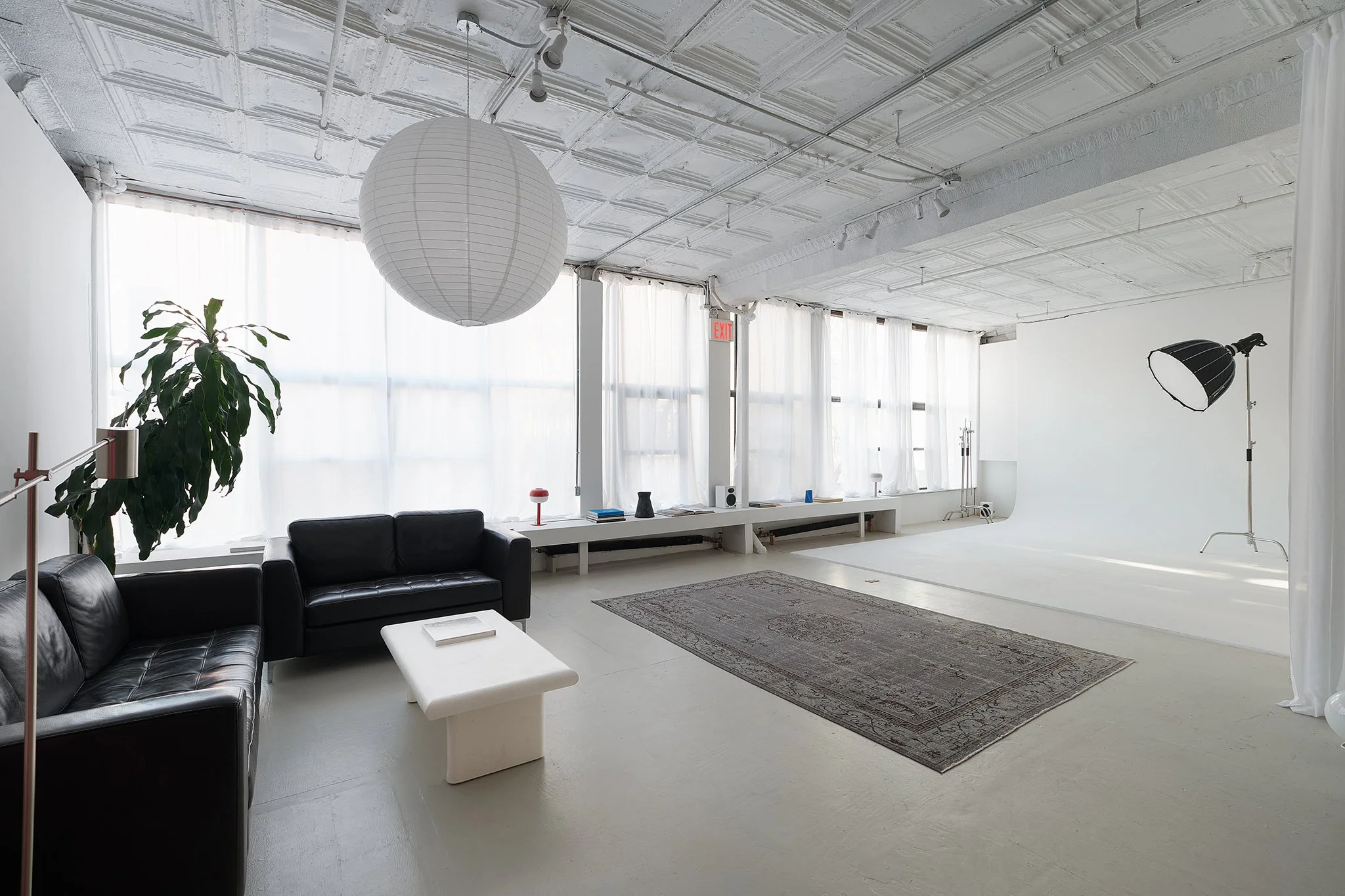 Bright studio space with a white ceiling, sheer curtains allowing natural light, black leather couches, a white ottoman, a potted plant, a decorative rug, a large paper lantern, and studio lighting equipment.