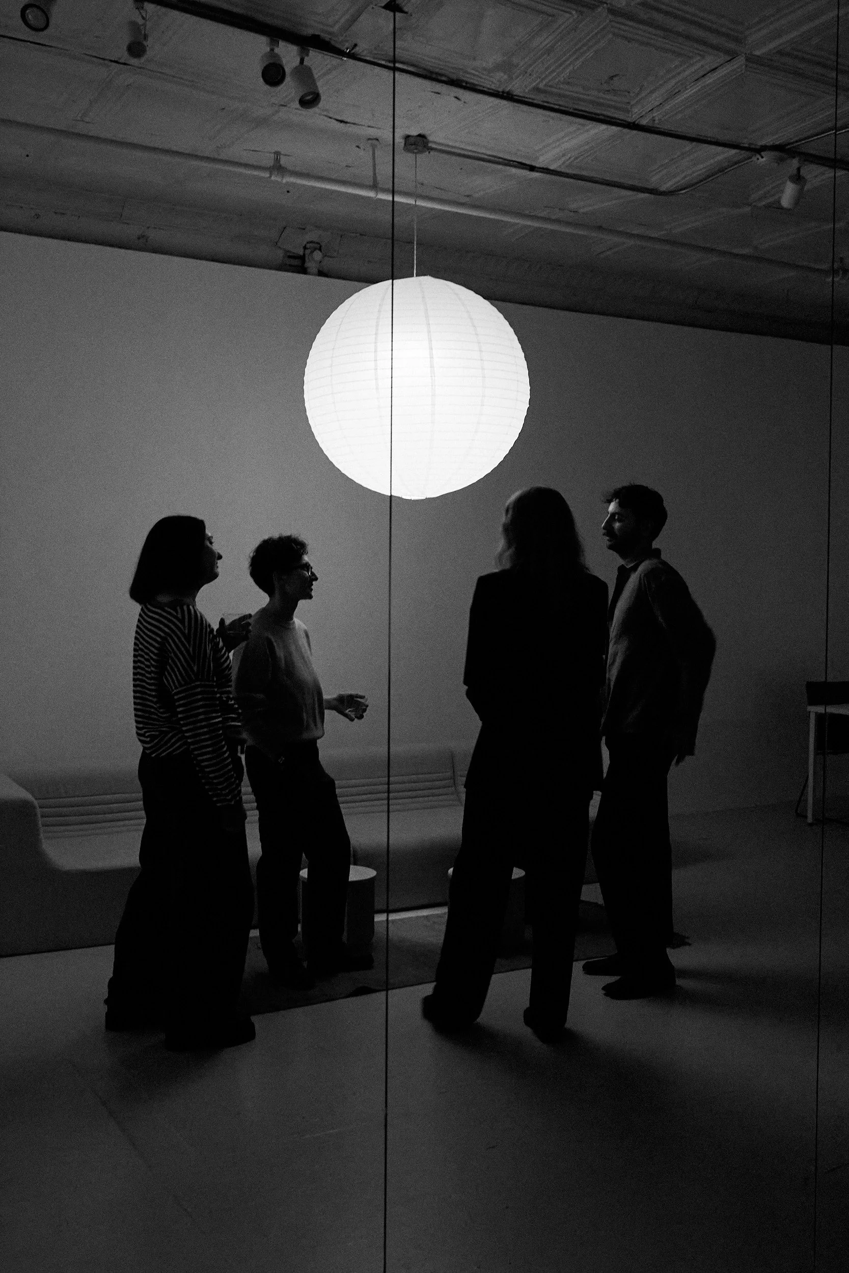 Four people in conversation are seen through a large mirror, with a lit spherical hanging lamp above them and a modern bench against the wall in the background.