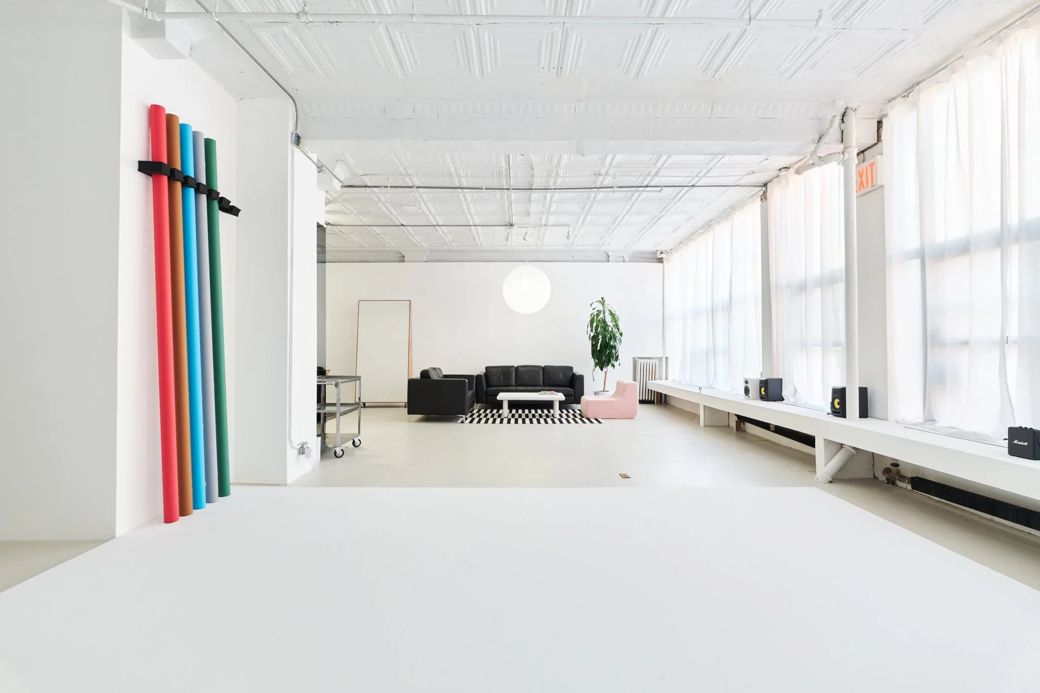 Bright spacious room with white walls and ceiling, large windows with sheer curtains, black couch, pink lounge chair, potted plant, and colorful foam tubes hanging on the wall.