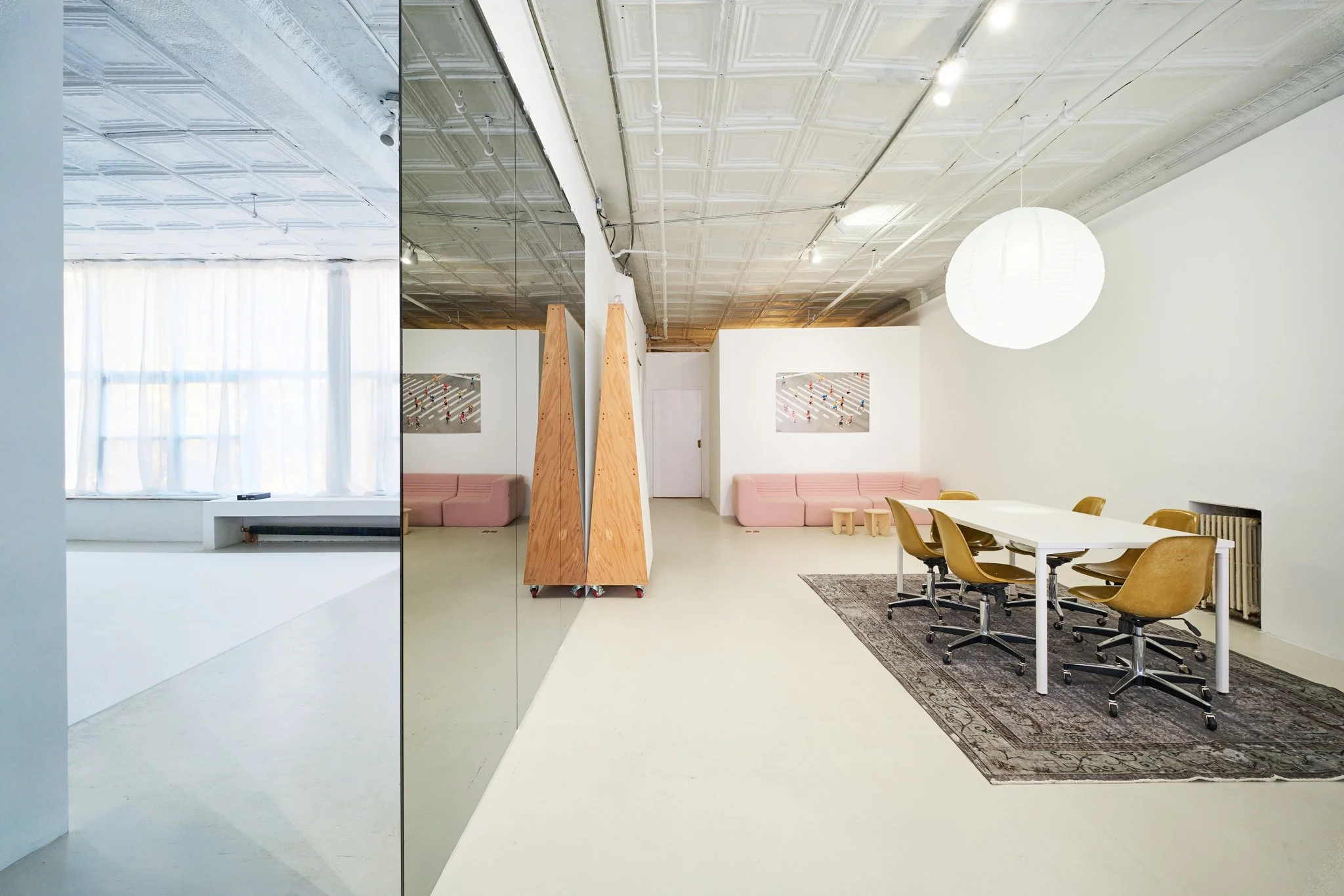 Modern office space with white walls, yellow chairs around a white table, pink couches, large window with curtains, artwork on the wall, and a large paper lantern light fixture.