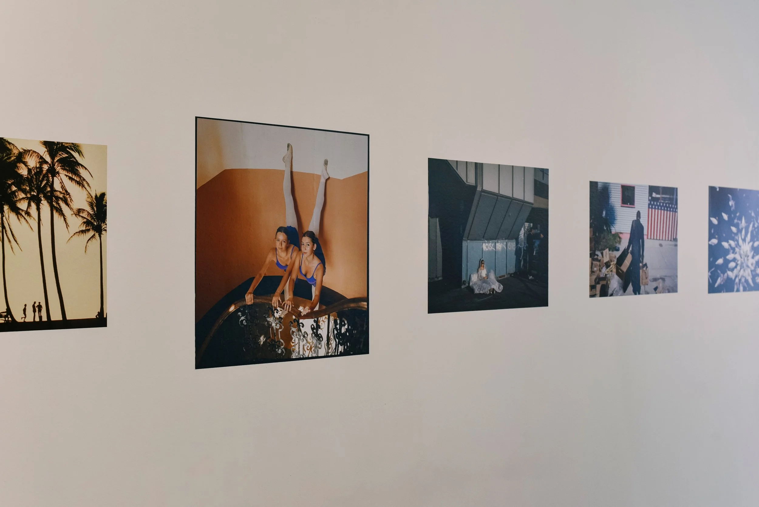 A series of five photographs displayed on a white wall. The photos include palm trees during sunset, two women in matching blue swimsuits with legs extended against an orange background, an industrial setting with a person sitting on the ground, a pe
