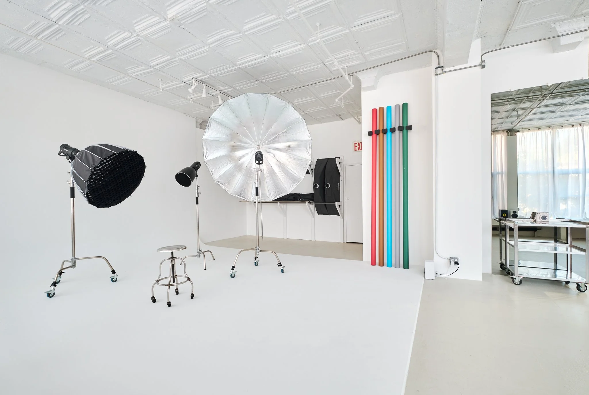 Photography studio with softbox, umbrella, and lights on a white backdrop, colorful rods on the wall, and a window with curtains.