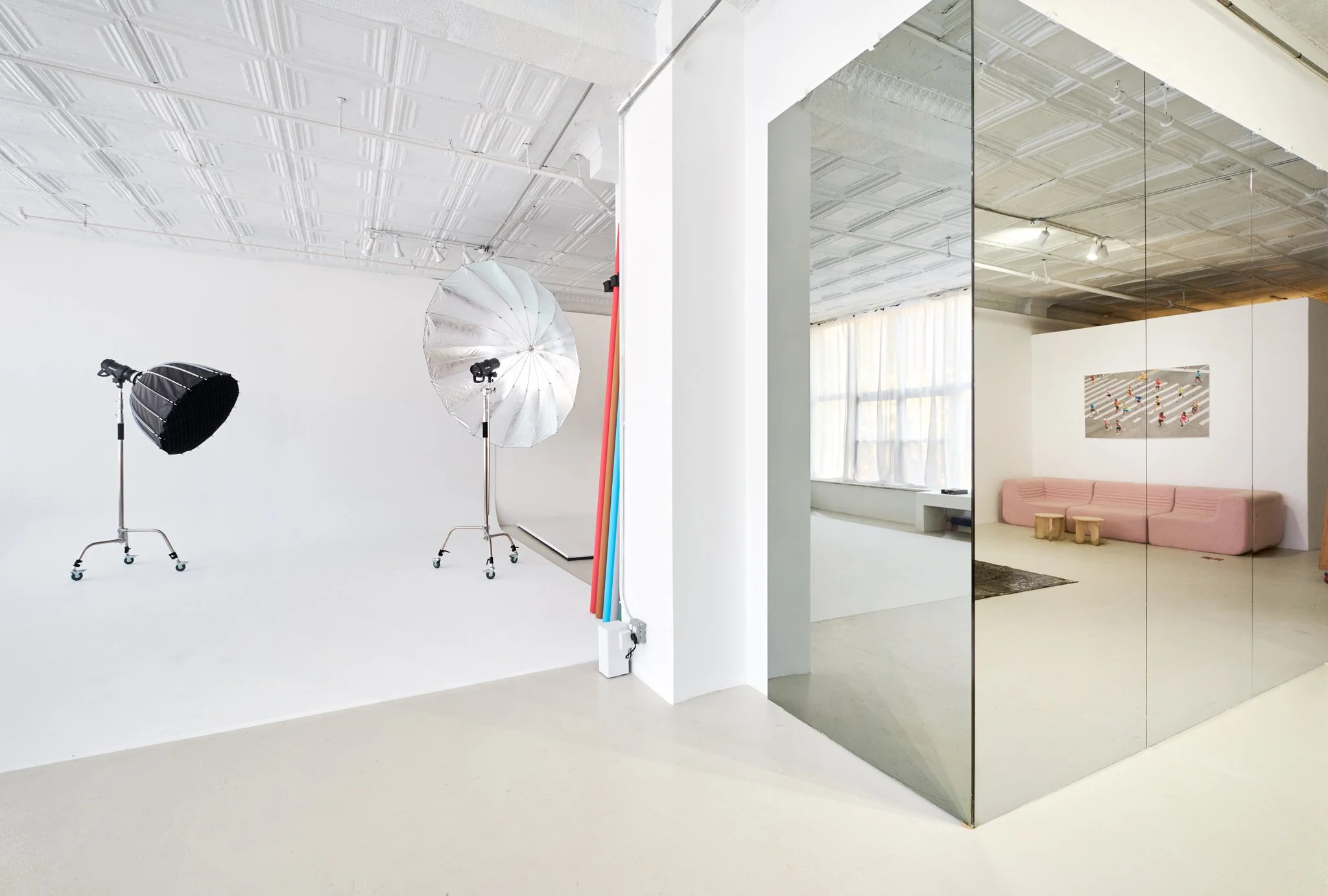 A photography studio with two light stands and umbrellas, white background, next to a mirror a room with a pink sofa, small stools, and wall art, large windows with curtains.