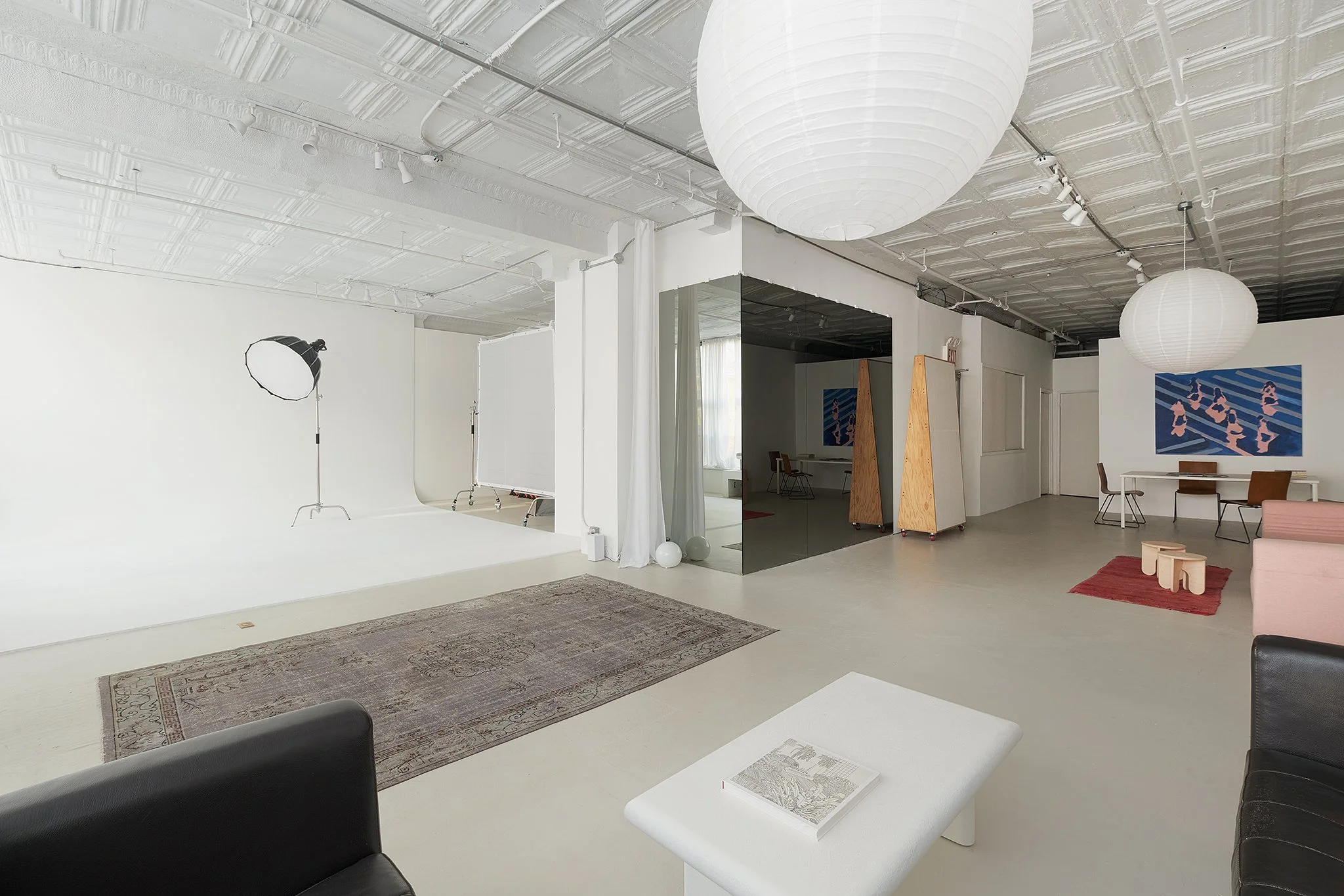 Interior of a modern, minimalist art studio with white walls and ceiling, featuring large paper lantern light fixtures, a photography studio setup with light and a white background, a couch, and artwork on the walls.