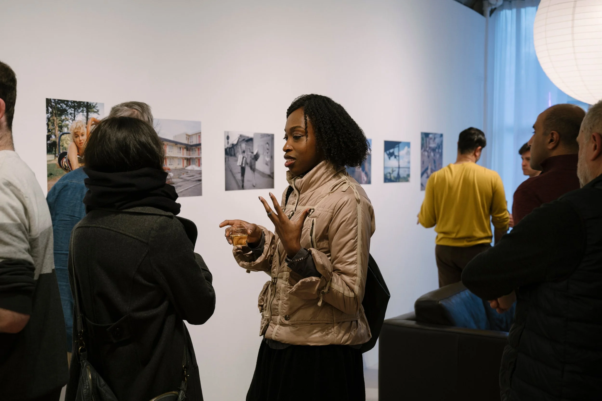 A group of people are gathered in an art gallery, looking at photographs on a white wall. A woman in a tan jacket is speaking to a woman in a black coat while holding a glass of wine.