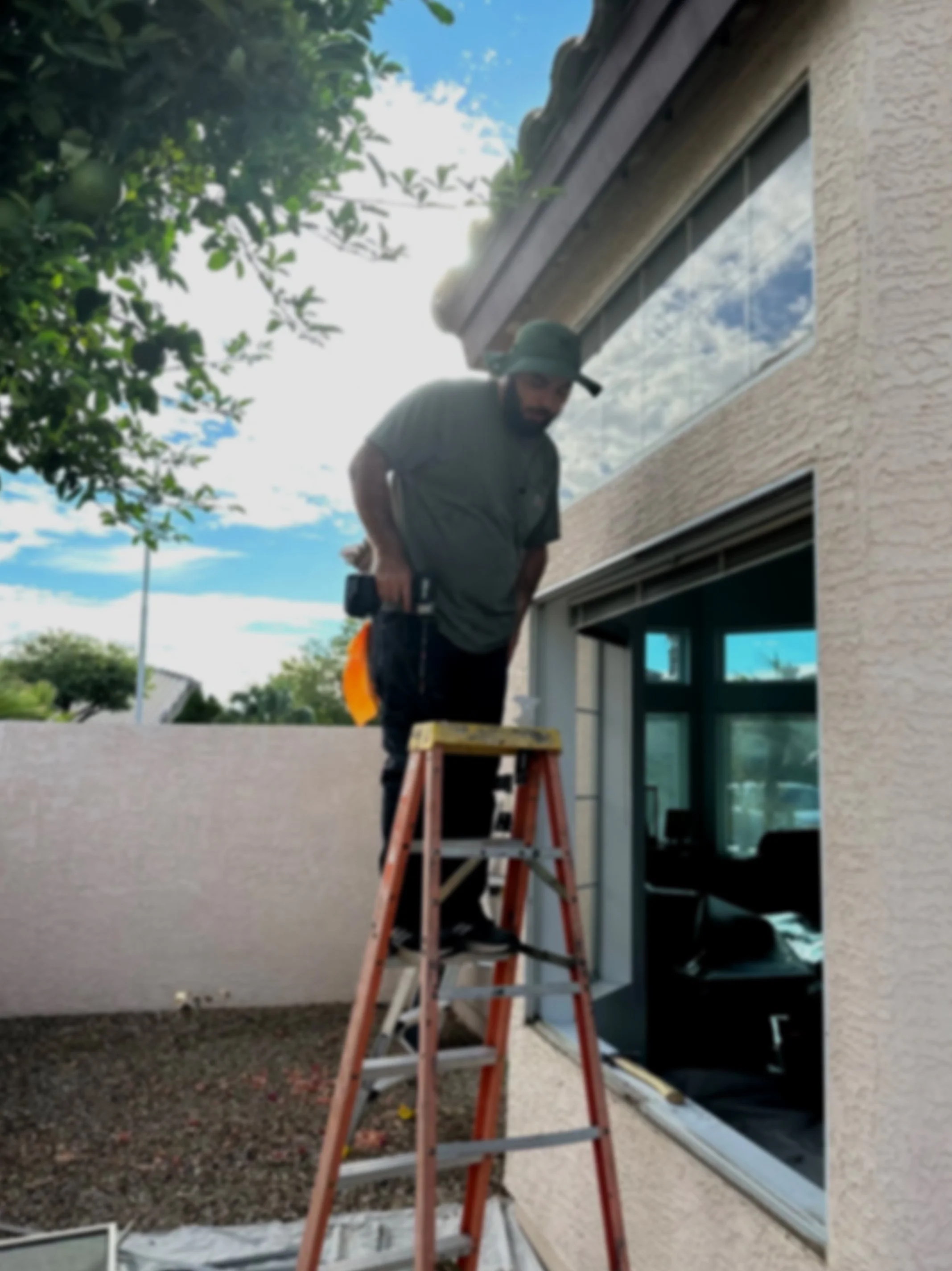 A man standing on a ladder outside a house, working on the window or wall, holding a tool, with trees and a partly cloudy sky in the background.