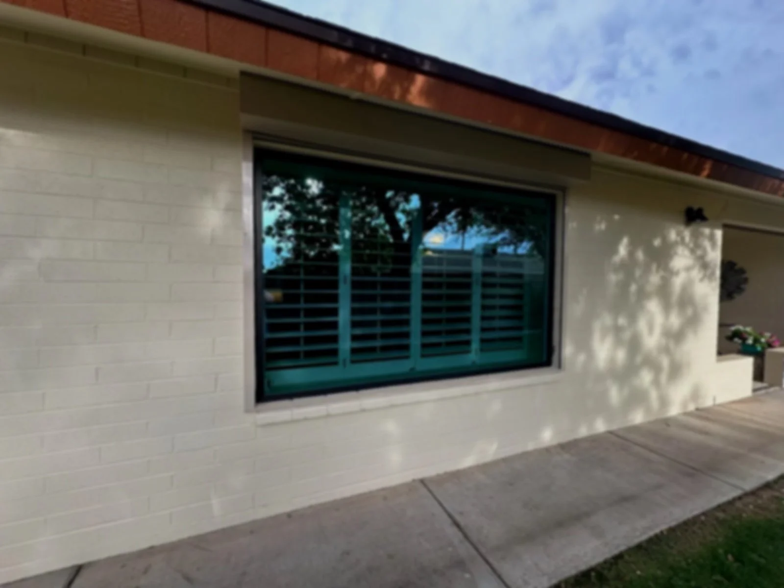 A house with a beige brick exterior, a large window with teal shutters, a sidewalk in front, and some flowers in a planter to the right.