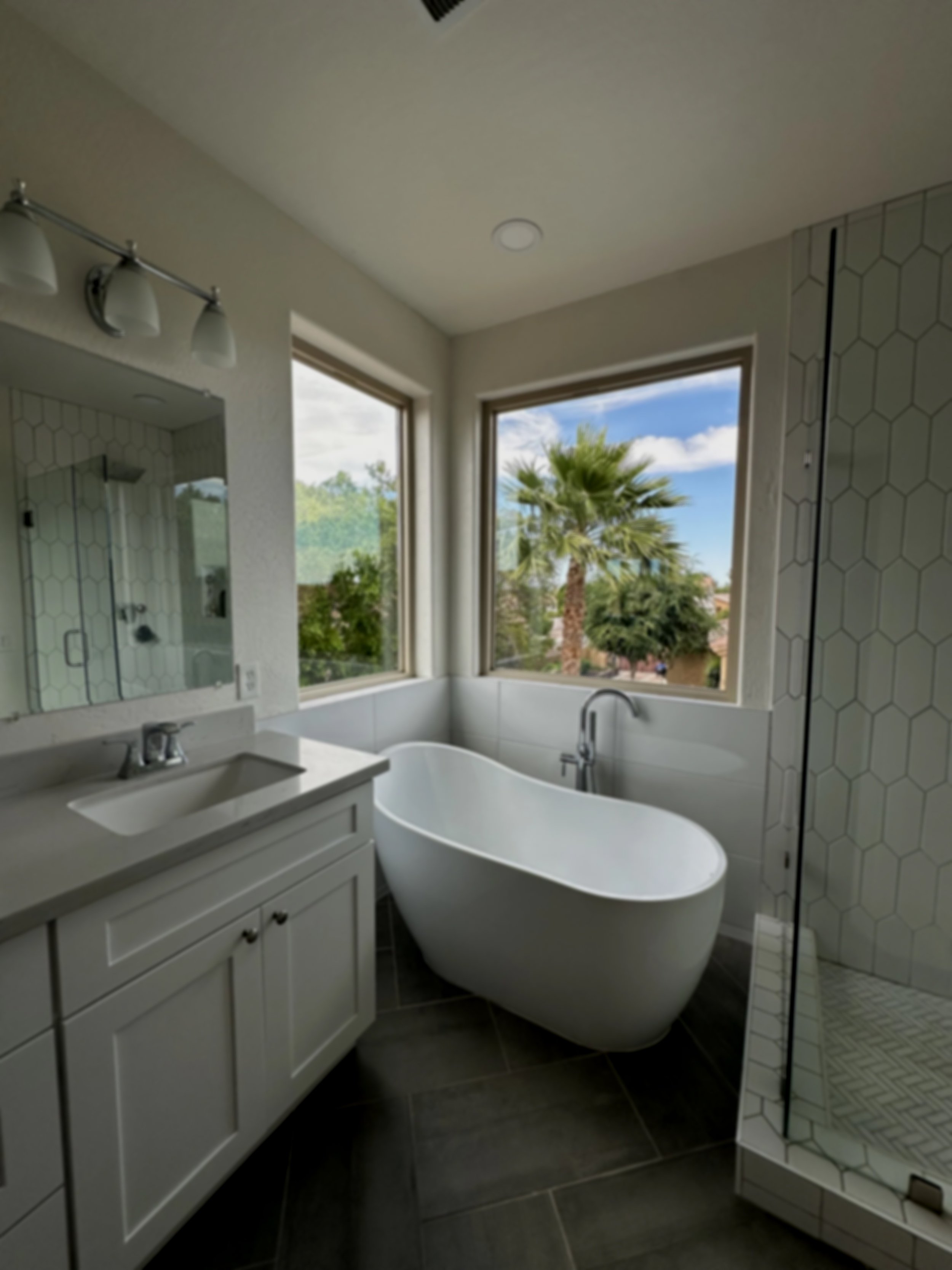 Modern bathroom with freestanding bathtub, large windows showing a palm tree outside, and a walk-in shower with tile walls.