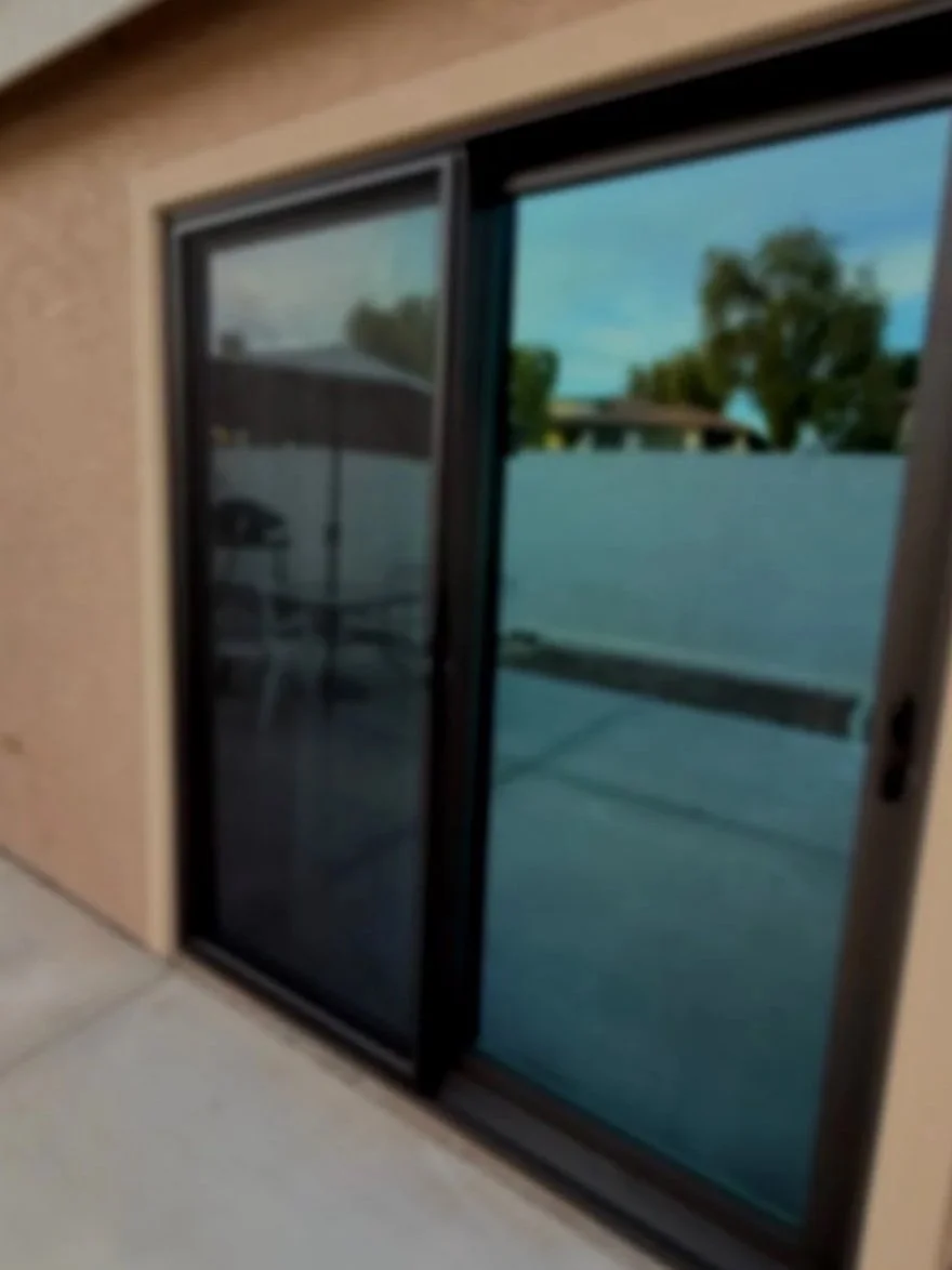 Exterior view of a sliding glass door with a security screen, leading to a backyard patio with trees and a building in the background.