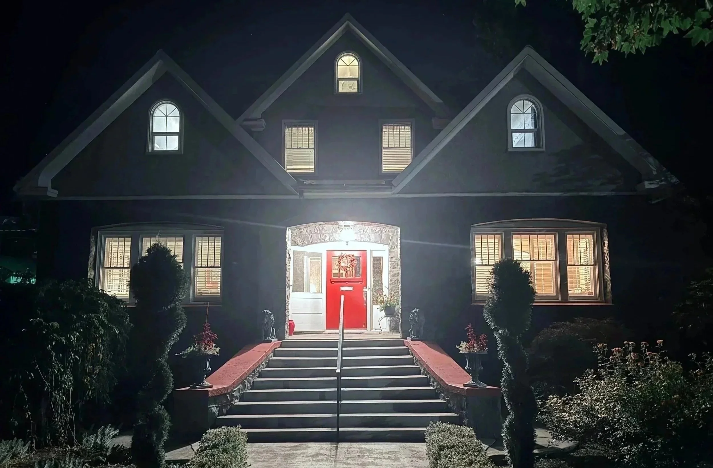 A two-story house at night with illuminated windows, a bright red front door decorated with a wreath, stone steps leading up to the entrance, and surrounding greenery.