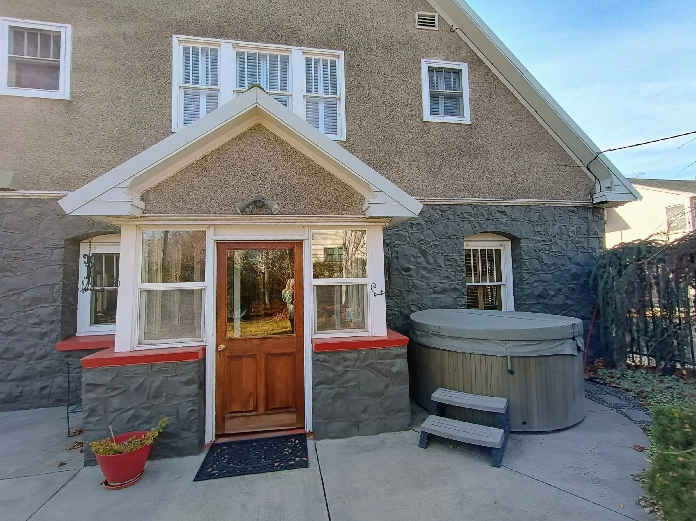 Backyard of a house with a small porch, hot tub, potted plant, and a stone wall, under a blue sky.