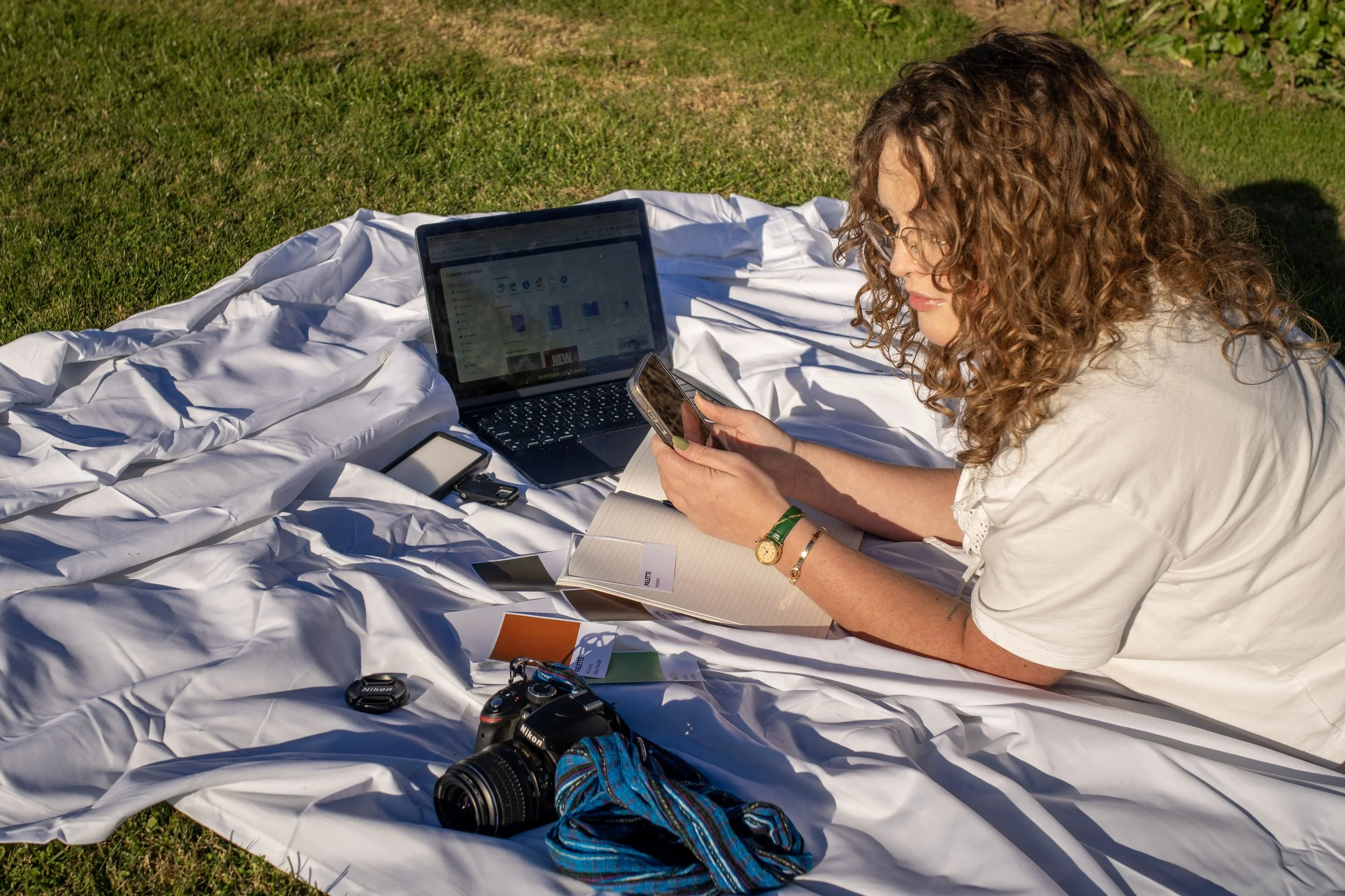 A digital marketer and social media manager woman with curly hair and glasses sitting on a white cloth outdoors on grass, working with a laptop, smartphone, camera, and notebooks. She is a content strategist for health and wellness brands.