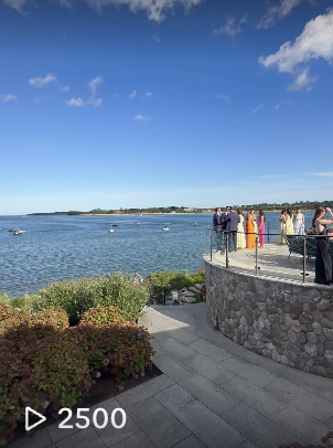 People gathering on a stone patio overlooking a body of water on a clear day with a few clouds in the sky.