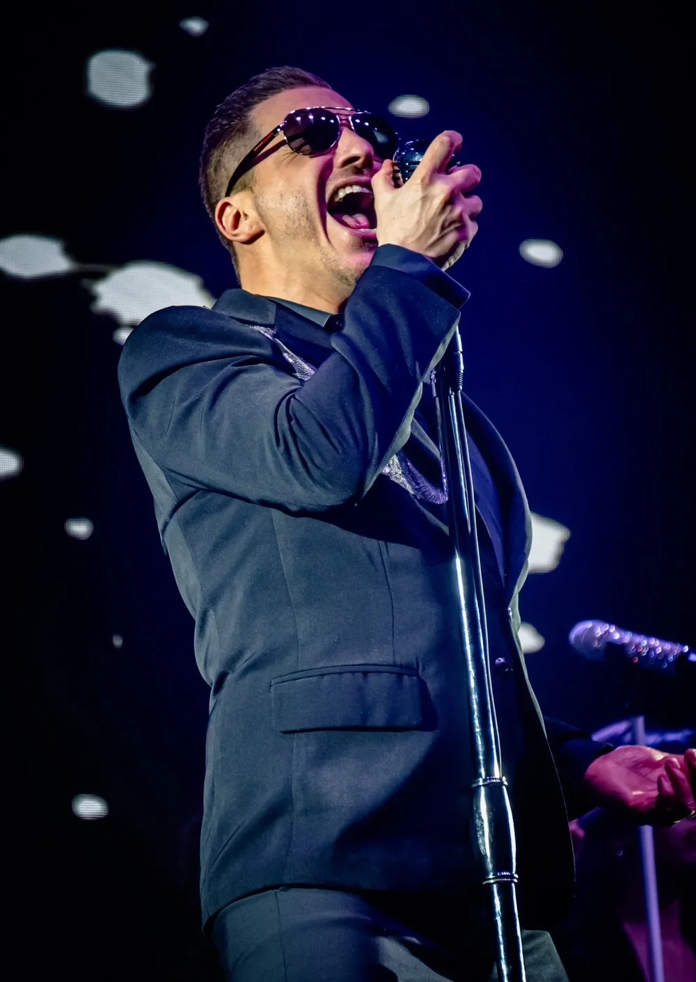 A male singer wearing sunglasses in a black suit singing into a microphone on stage.