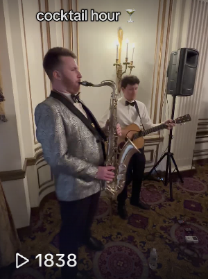 Two musicians playing instruments at a cocktail hour event, one on saxophone and the other on guitar, in an elegant room with wall sconces and a speaker.