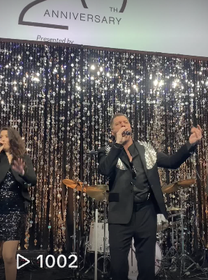 A man and a woman singing on stage during a celebration event with a shiny, sequin curtain backdrop.