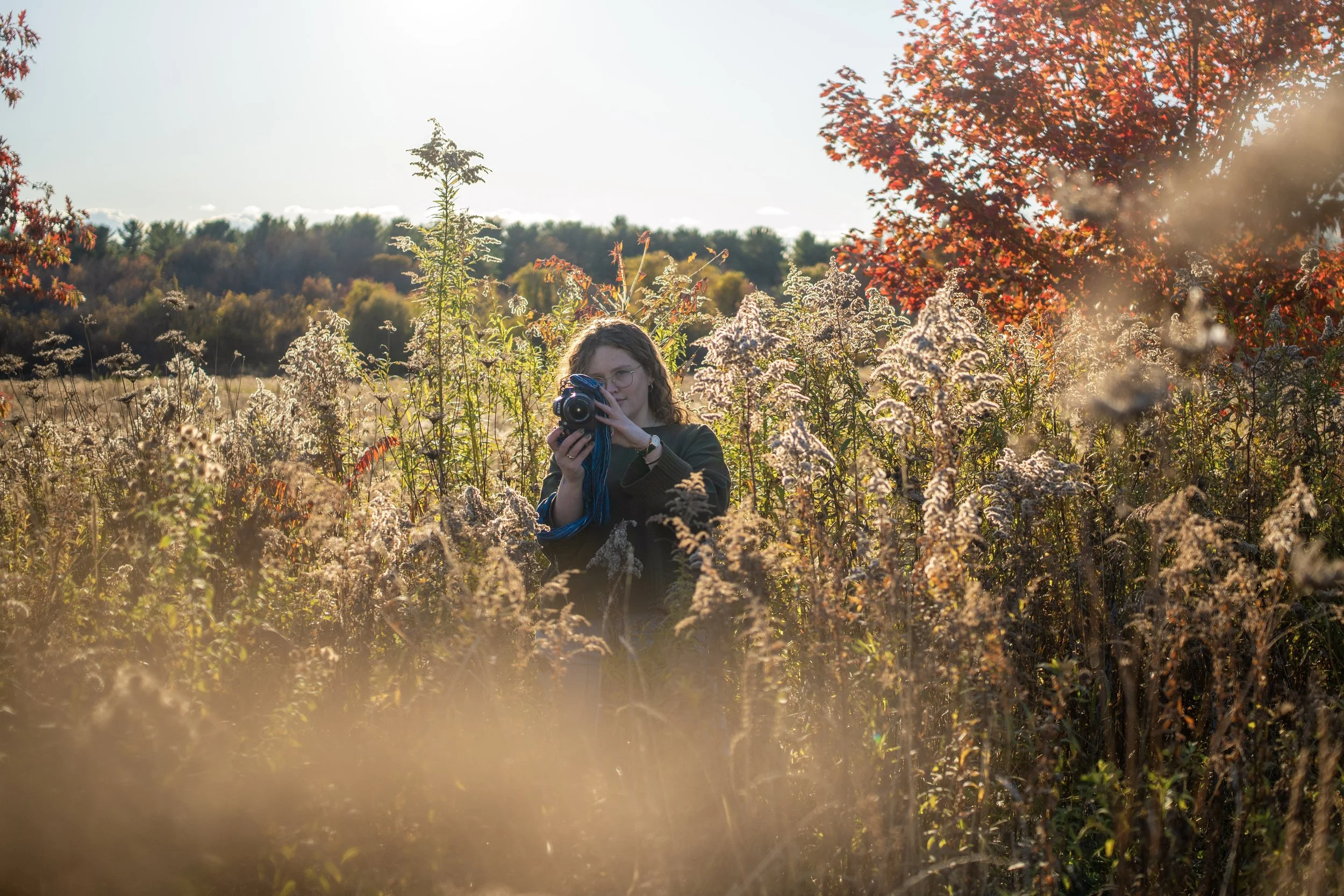 A woman with glasses taking a photograph with a camera in a field of tall, dried flowers during sunset, with trees and a red-leafed tree in the background.