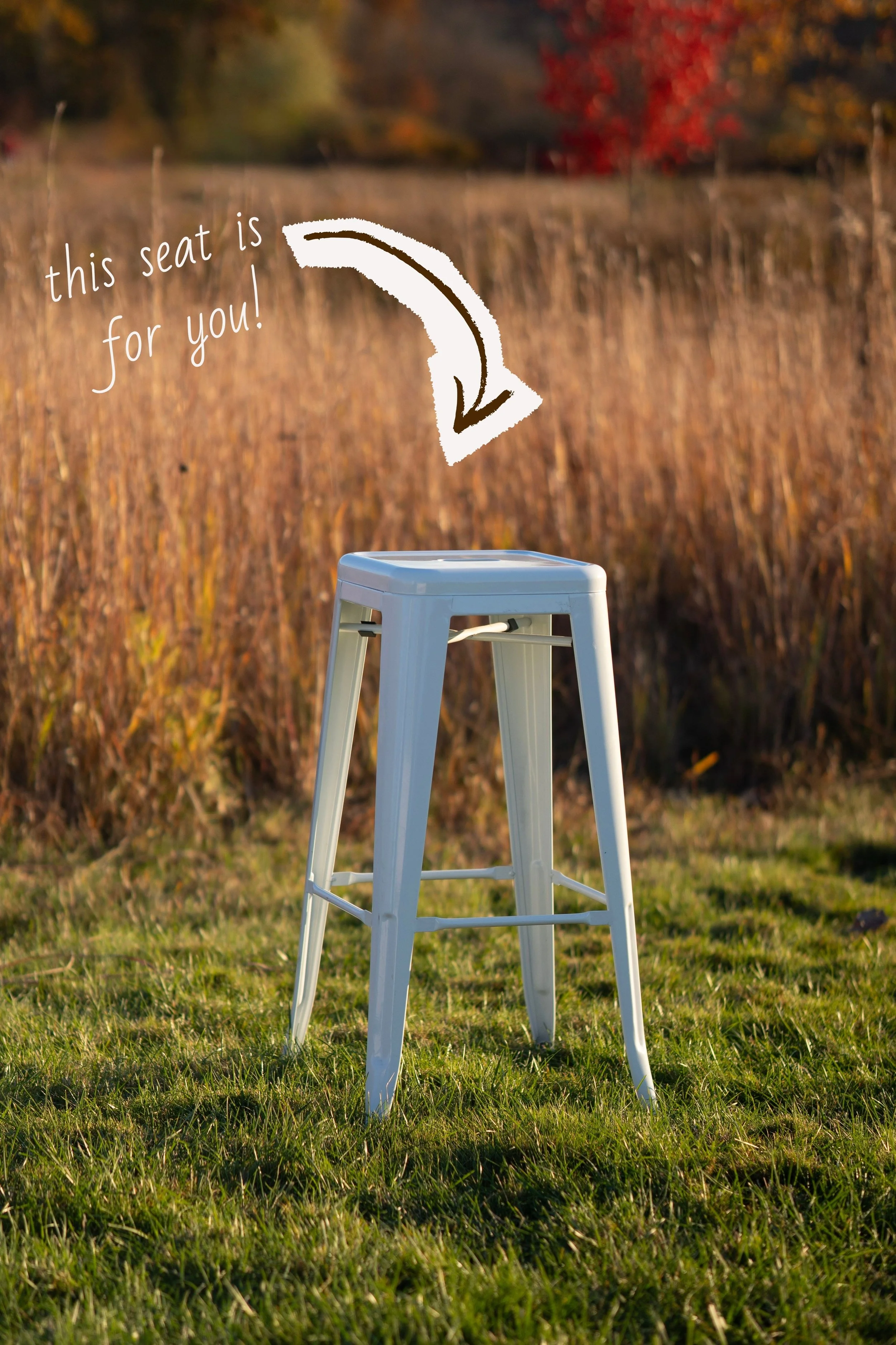 A white outdoor bar stool on green grass, with a blurred autumnal background of trees with fall foliage, and a handwritten message pointing to the stool that says "this seat is for you!".