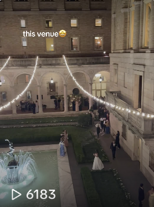 Evening event with string lights across courtyard, fountain, and guests in formal attire outside large building.