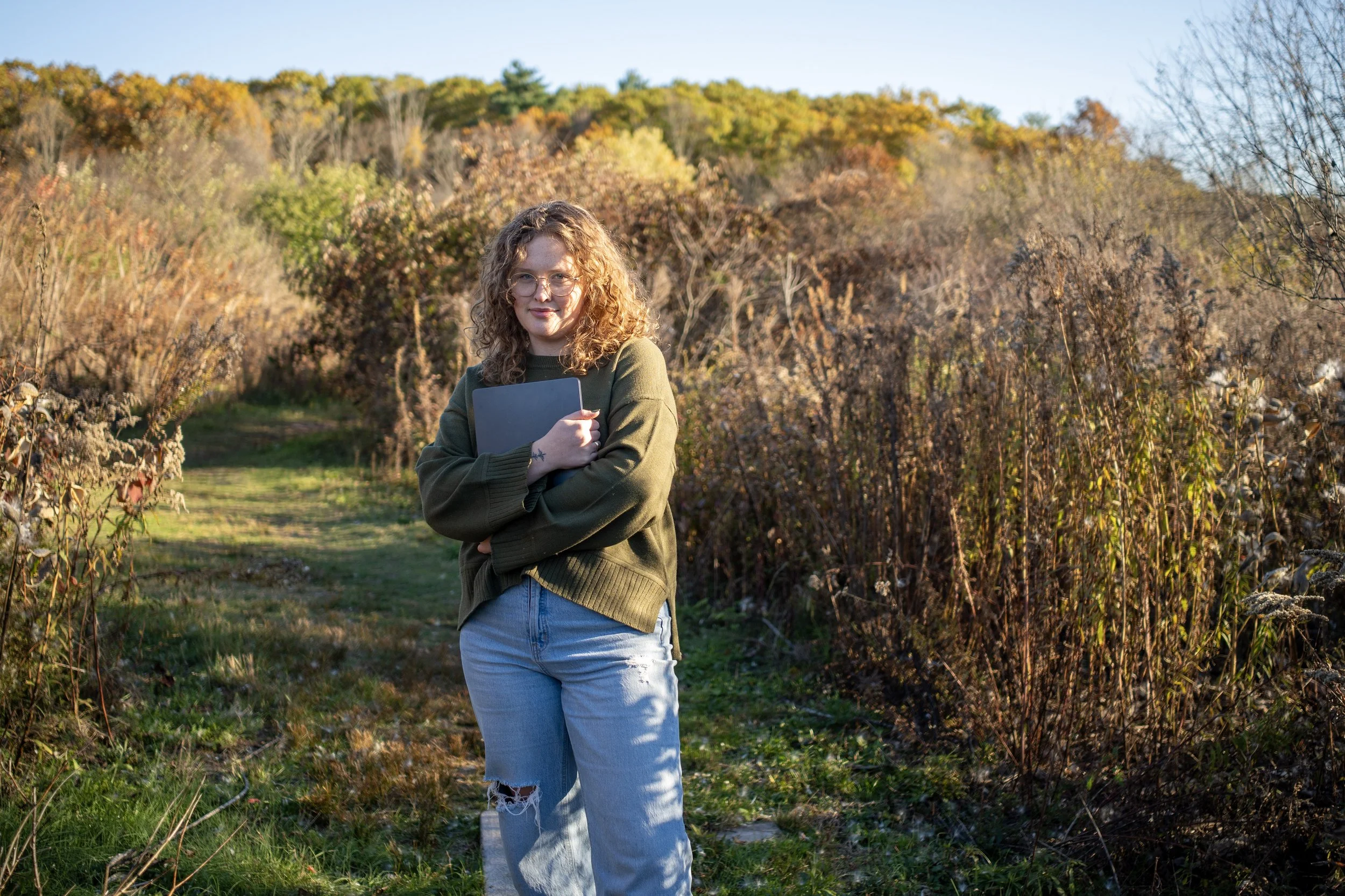 A social media manager and digital marketer woman with curly hair and glasses standing on a grassy path in an autumn landscape, holding a laptop and smiling. She is a content strategist for health and wellness brands.