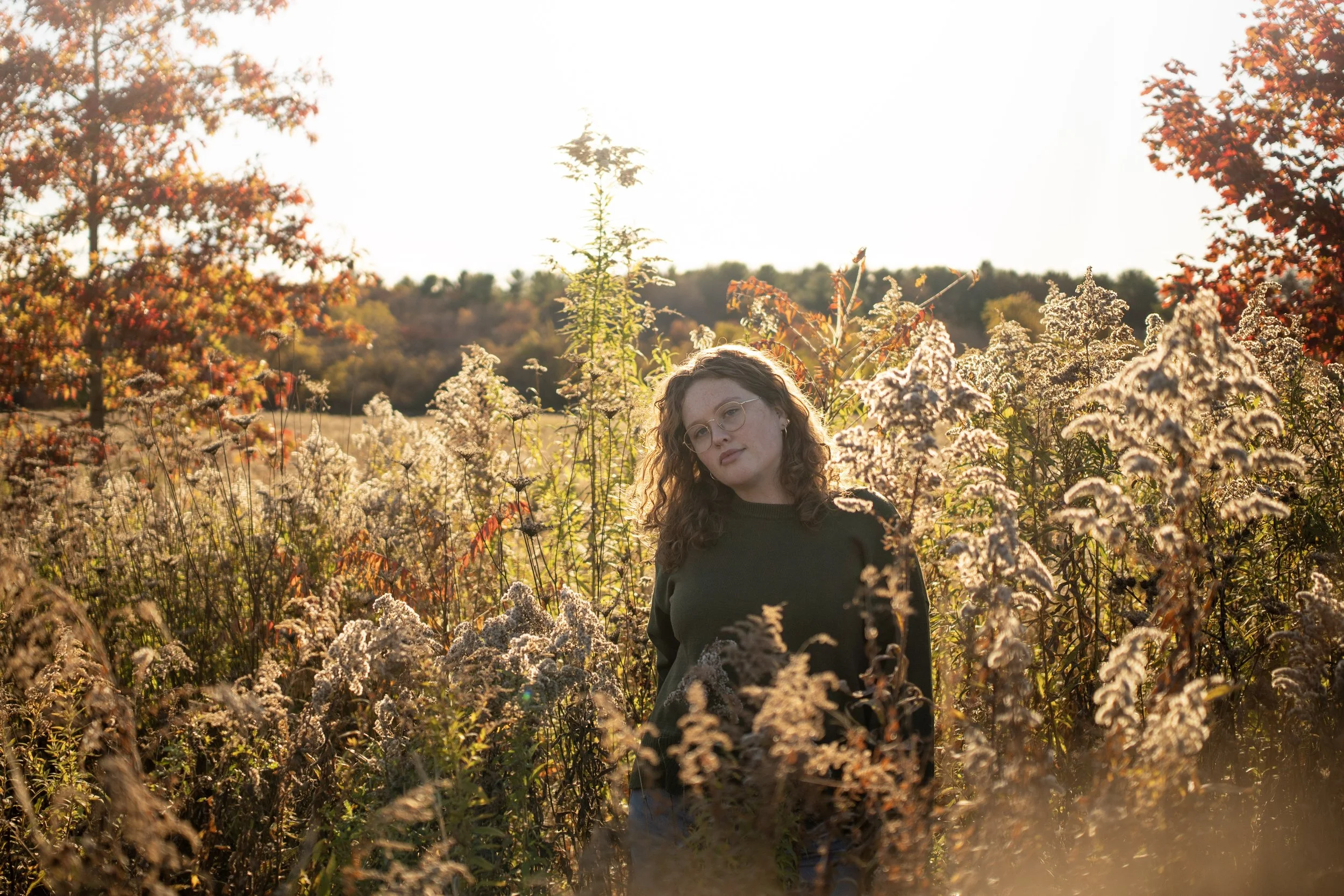 A young woman with curly hair and glasses stands in a field of tall, flowering plants during sunset. She is wearing a dark sweater and looks contemplative amidst the autumn scenery. She is a content strategist and social media manager.