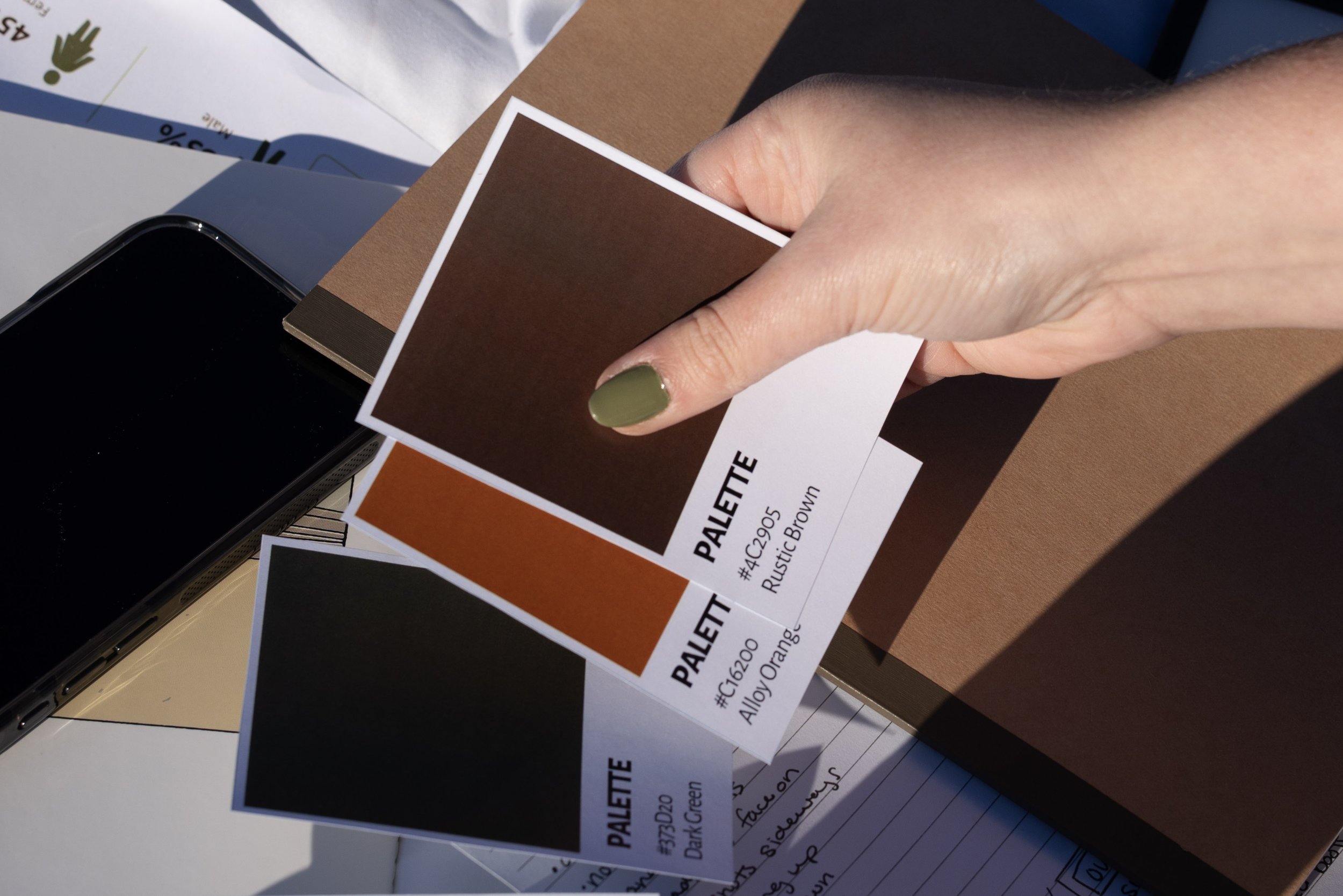 A hand holding paint color samples labeled 'PALETTE' in shades of brown, orange, and black. The sample in focus is 'Rustic Brown' and 'Ally Orange'.
