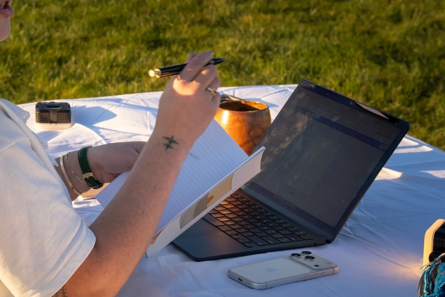 Social media manger, content strategist, and digital marketer writing in a notebook outdoors with a laptop, smartphone, and a copper mug on a white tablecloth in a grassy area during daylight