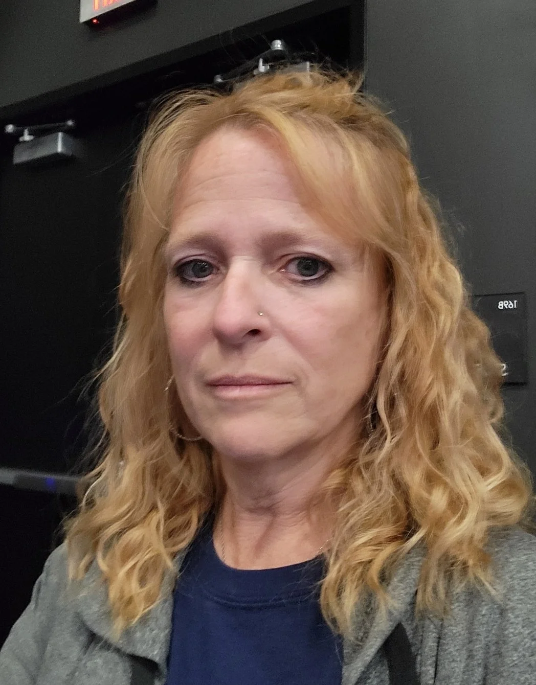 Close-up of a woman with wavy, shoulder-length blonde hair, light skin, wearing a gray jacket over a navy blue top, standing indoors with dark walls and electronic equipment in the background.
