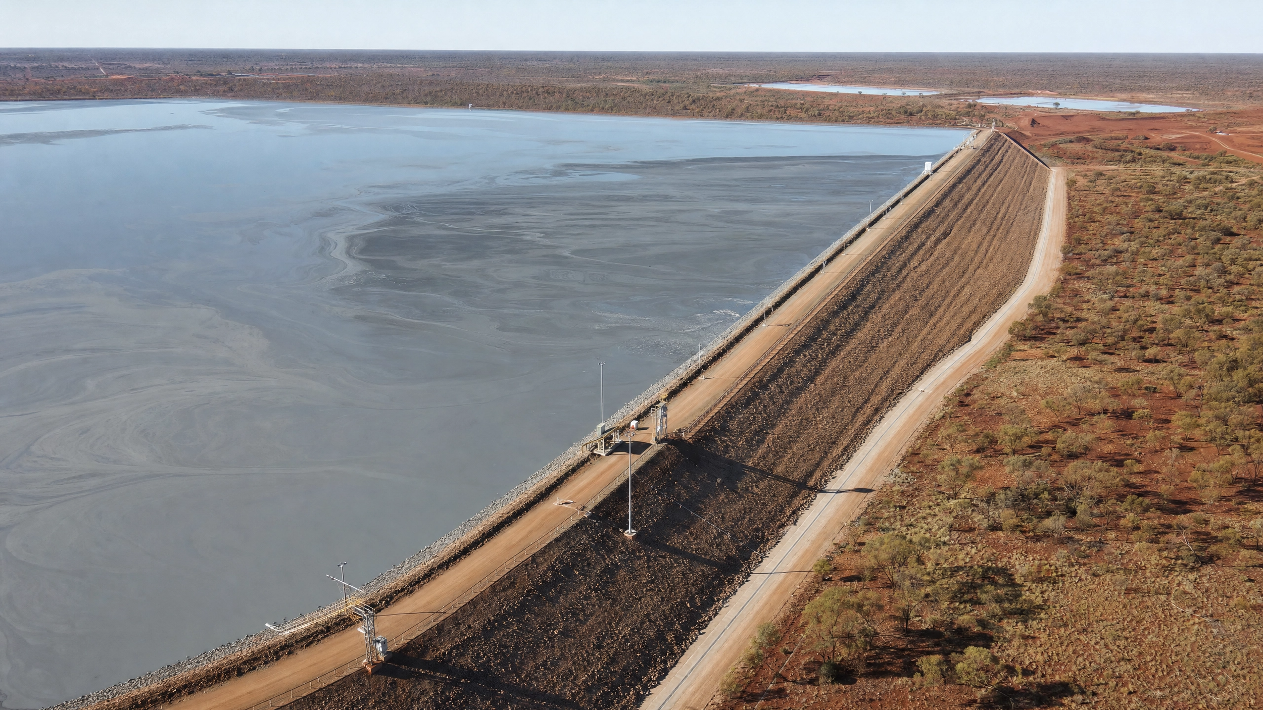 A large dam holding back water in an arid landscape with sparse trees and a dirt road along the top of the dam for tailing dam inspections by DeepSky IQ.